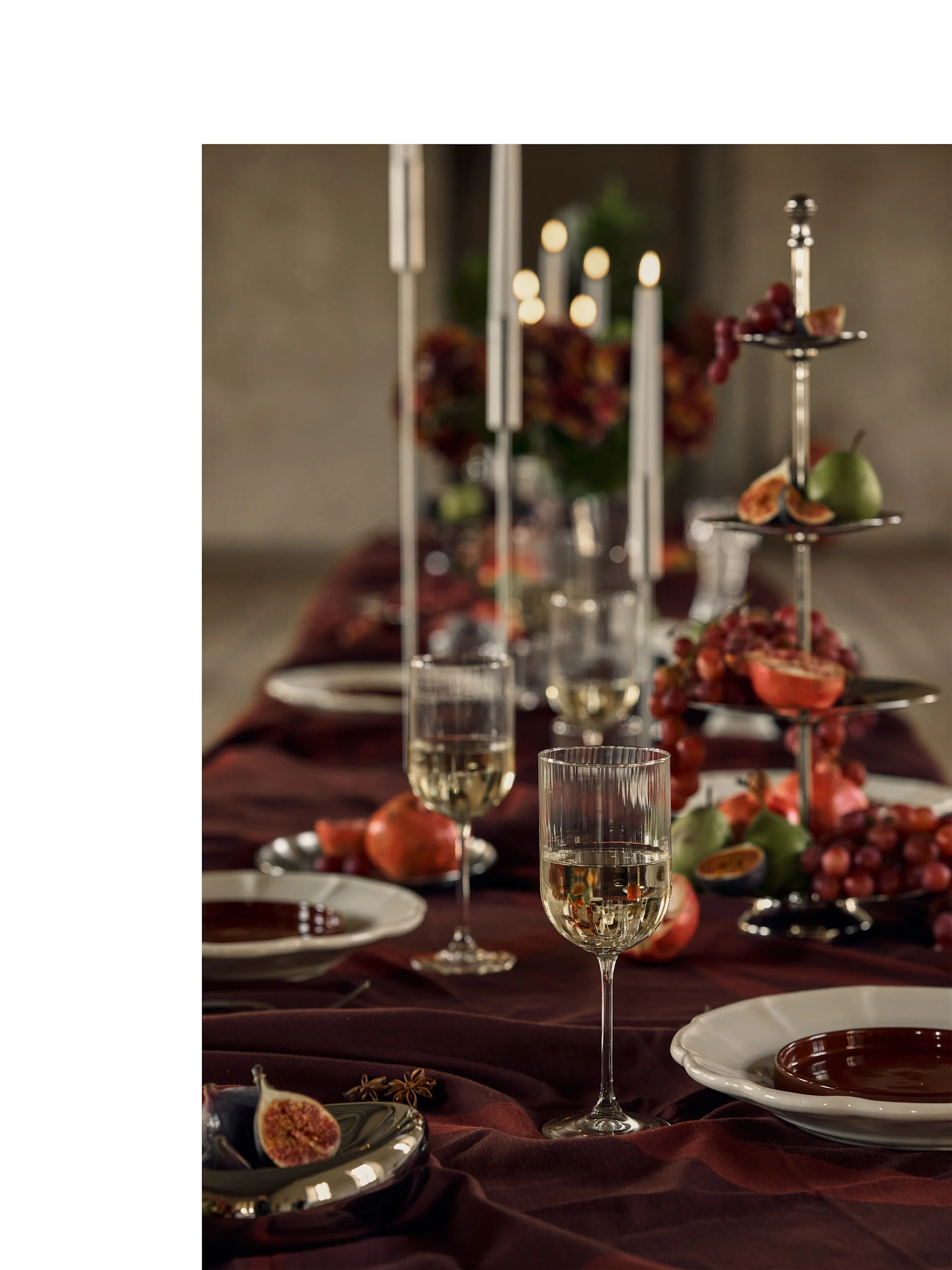 Feestelijke tafel gedekt met witte wijnglazen, borden en een etagère gevuld met kleurrijke vruchten en kaarslicht.