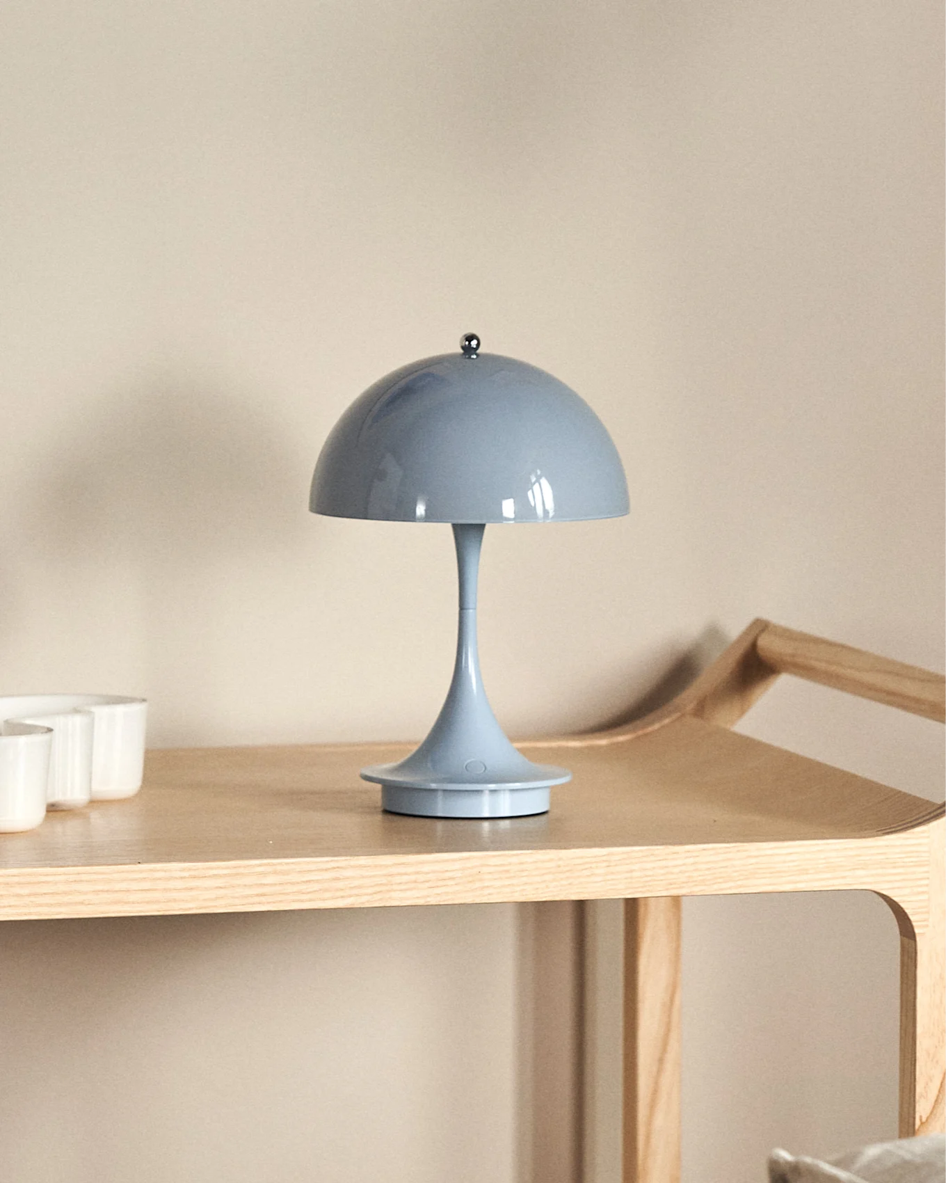 Een lichtblauwe paddenstoelvormige tafellamp op een lichte houten plank, met witte kopjes deels zichtbaar aan de linkerkant.