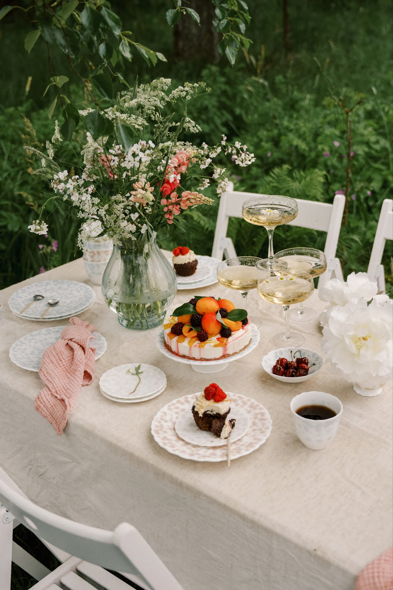 Buitentafel gedekt met bloemenvaas, champagne, fruittaart en cupcakes met bessen.