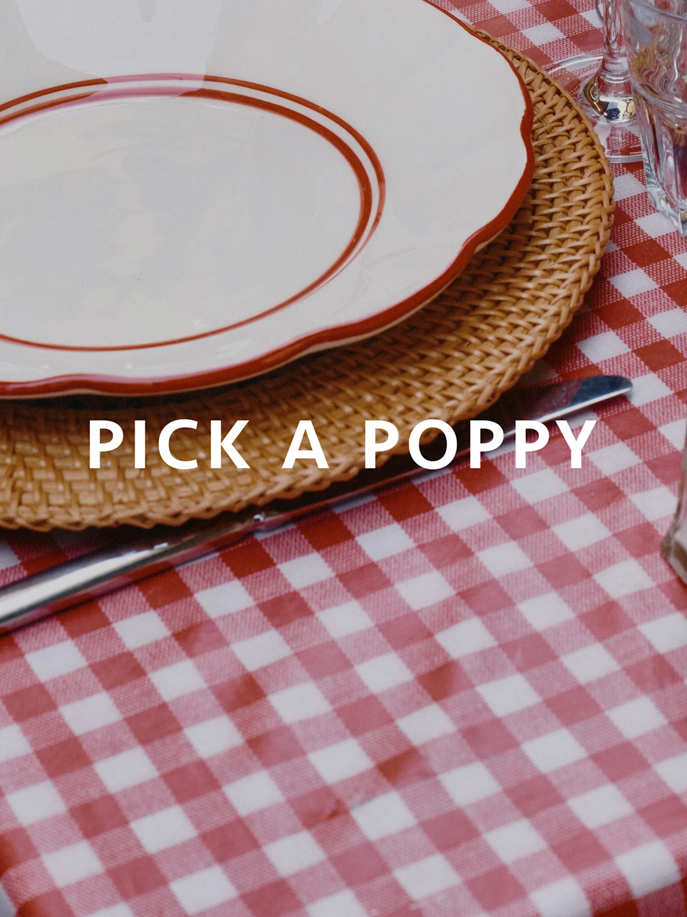 Gedekte tafel met rood-wit geblokt tafelkleed, gevlochten placemat en wit bord met rode rand. Tekst: PICK A POPPY.