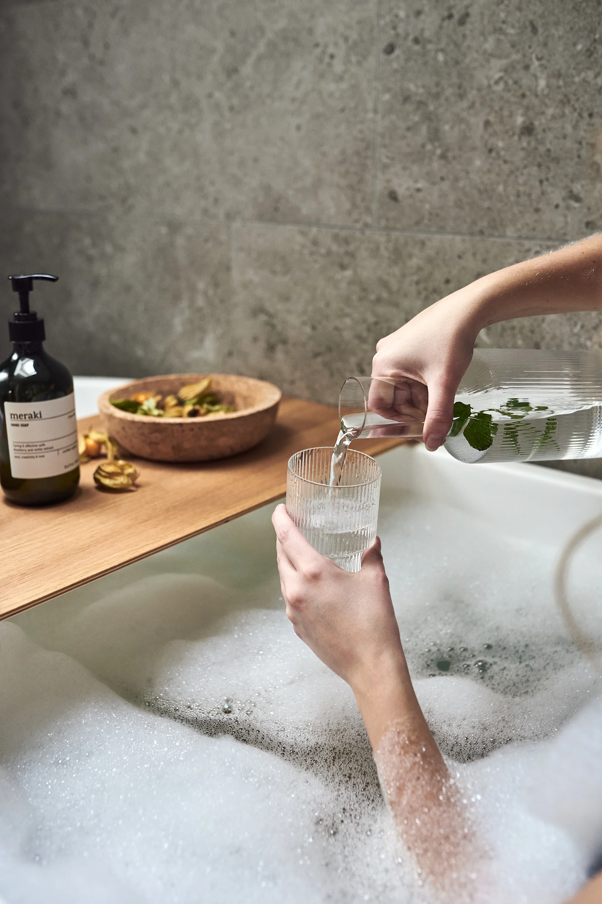 Handen gieten water in een ribbelglas van Ferm Living als onderdeel van een spa-avond thuis met een warm bubbelbad. 