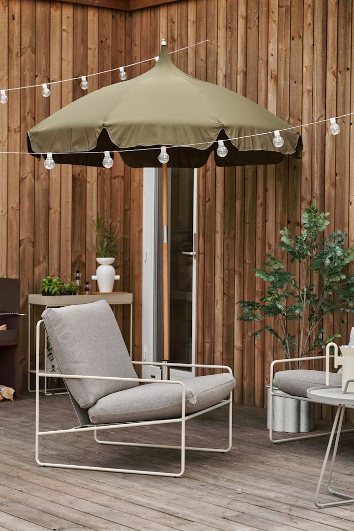Een gezellig houten terras met sprookjesachtige verlichting, Lull parasol en Desert loungestoel, beide van Ferm Living.