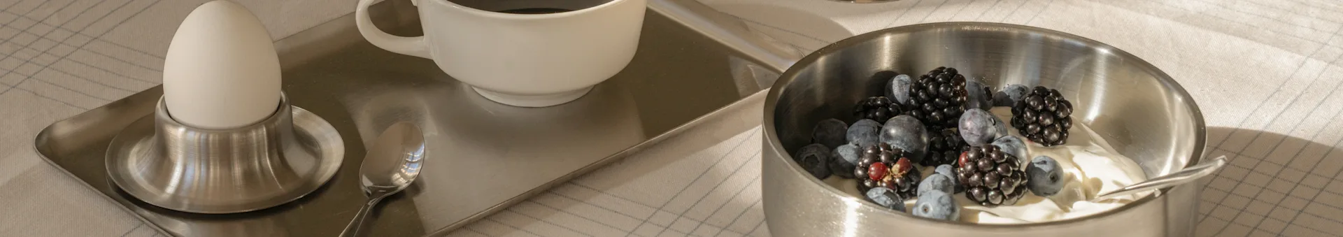 Ontbijtblad met een gekookt ei, koffie en een kom yoghurt met bosbessen en bramen op een geblokt tafelkleed.