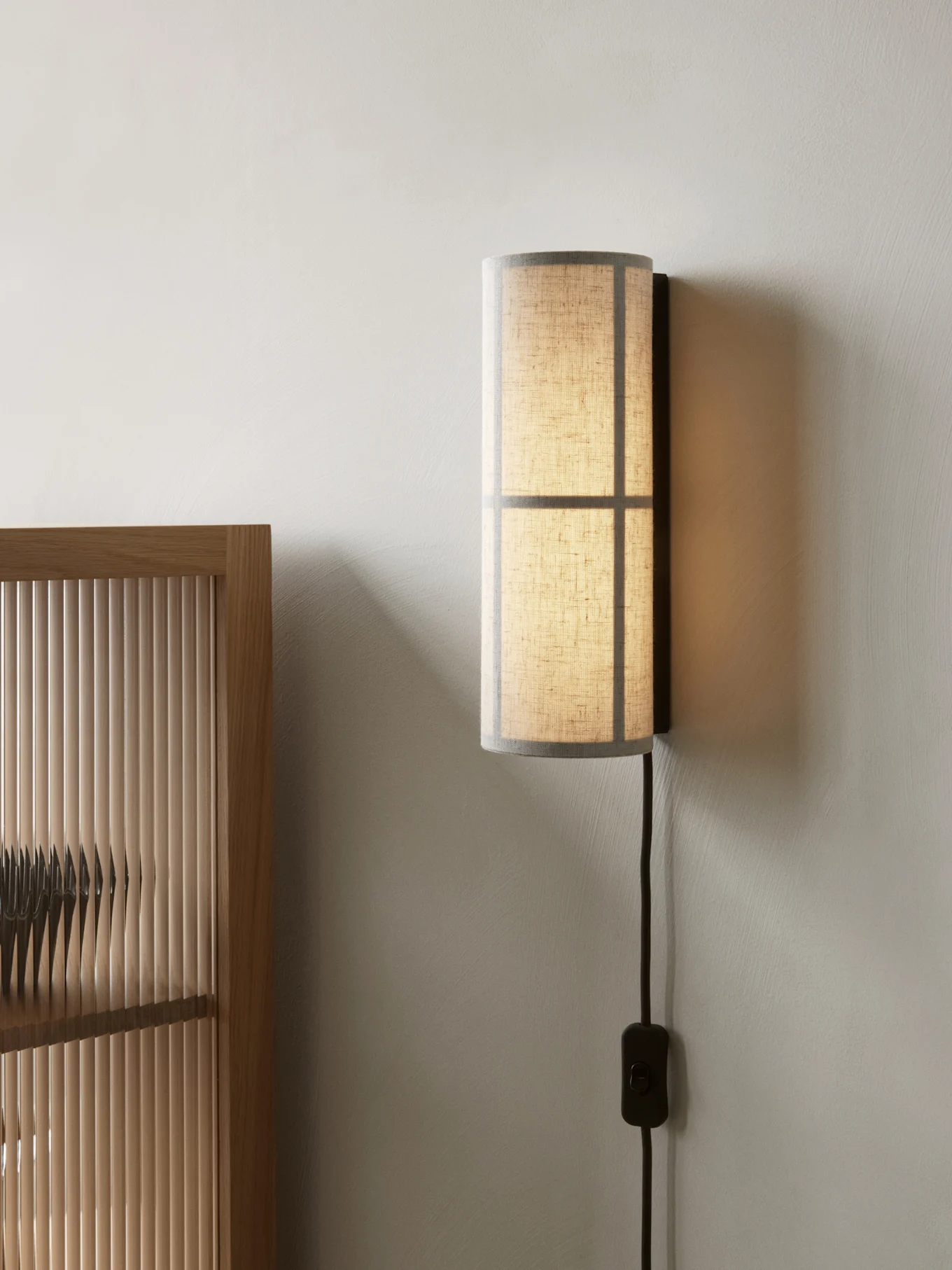 Cilindrische wandlamp met een doorschijnende stoffen kap, gemonteerd op een lichte muur naast een houten lattenwand.