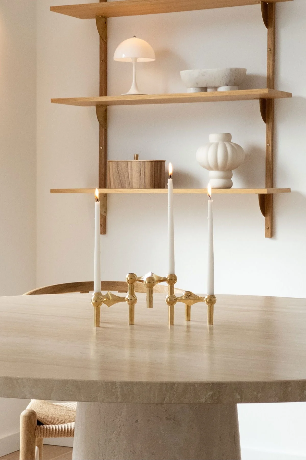 Een gouden kandelaar met drie brandende kaarsen op een ronde tafel, met planken en decoratieve objecten erachter.