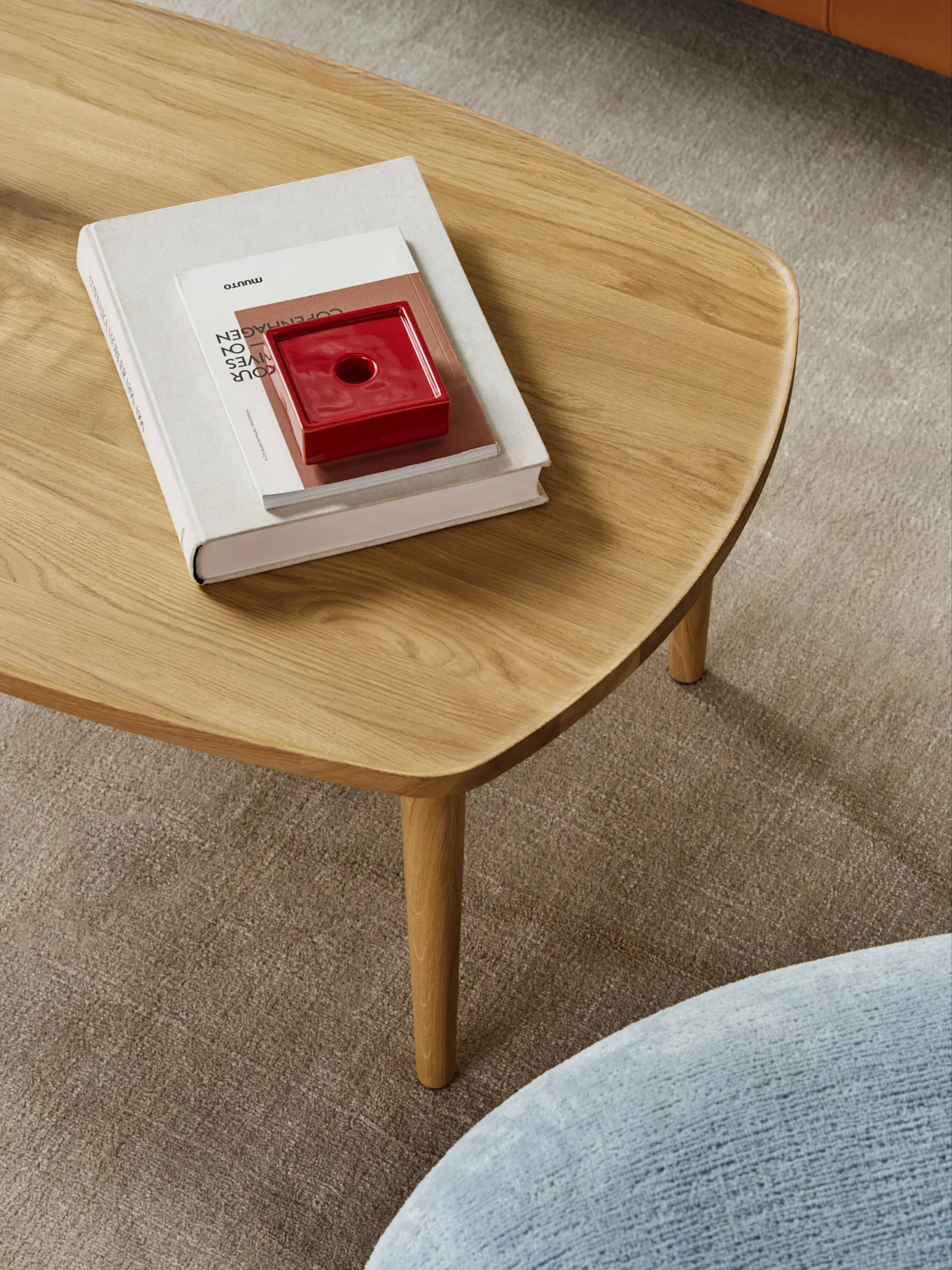 Een licht houten salontafel met afgeronde randen, met een stapel boeken en een rood keramisch object, op een beige tapijt.