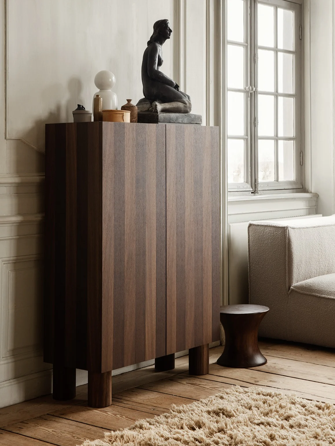 Donkere houten kast met verticale strepen, met daarop een zittend beeld, witte lamp en decoratie, naast een getextureerde bank en donkere kruk.