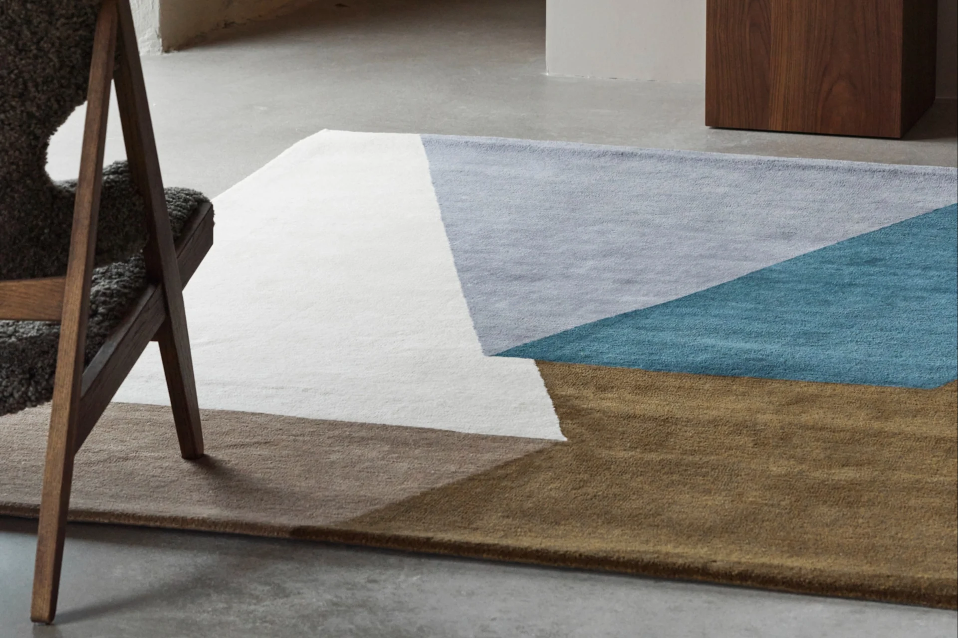 Een geometrisch vloerkleed in wit, lichtblauw, turquoise en bruin op een betonnen vloer, met een houten stoel en een blauwe vaas.