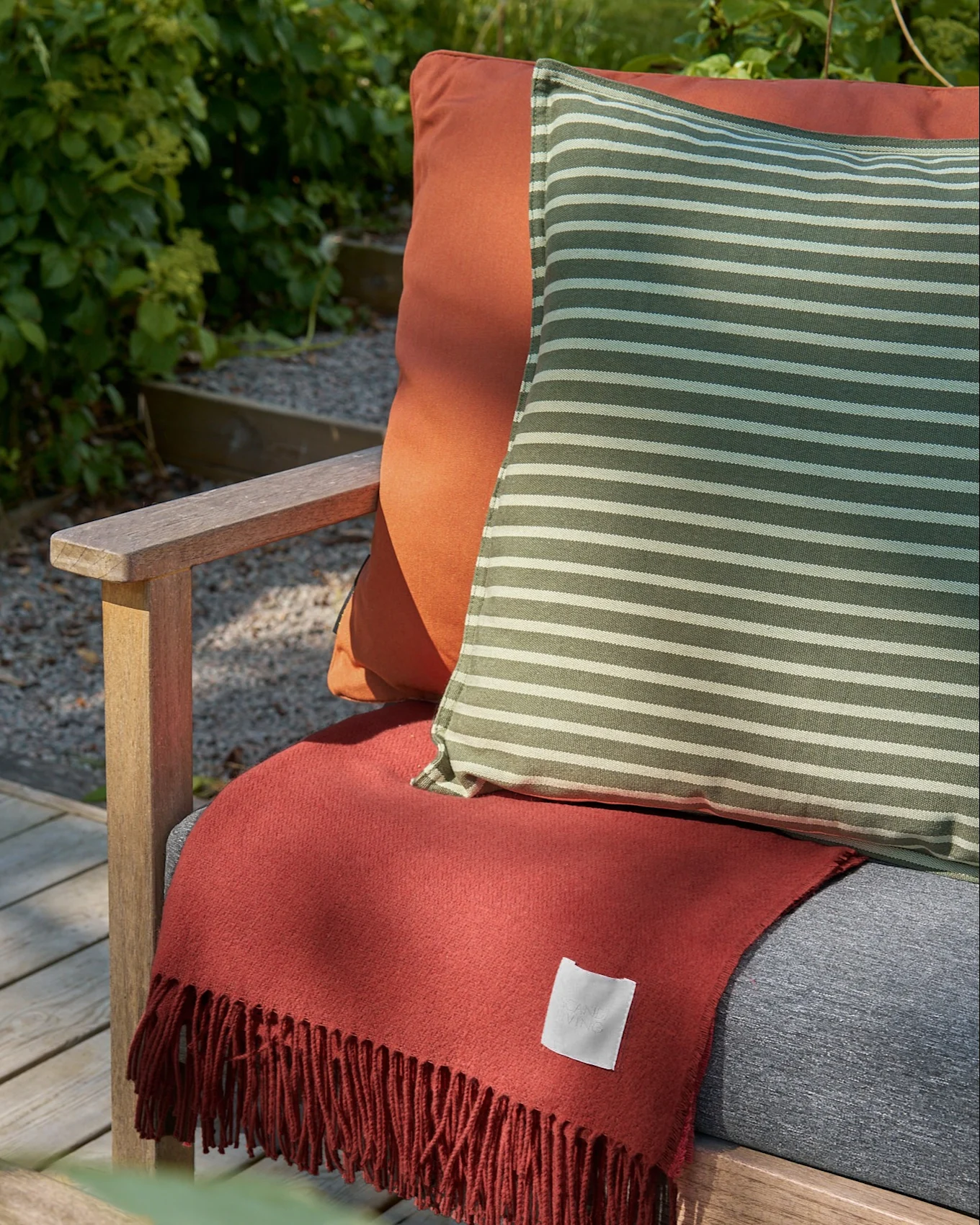 Buiten houten stoel met een oranje kussen, een gestreept groen-wit kussen en een terracotta franje deken.