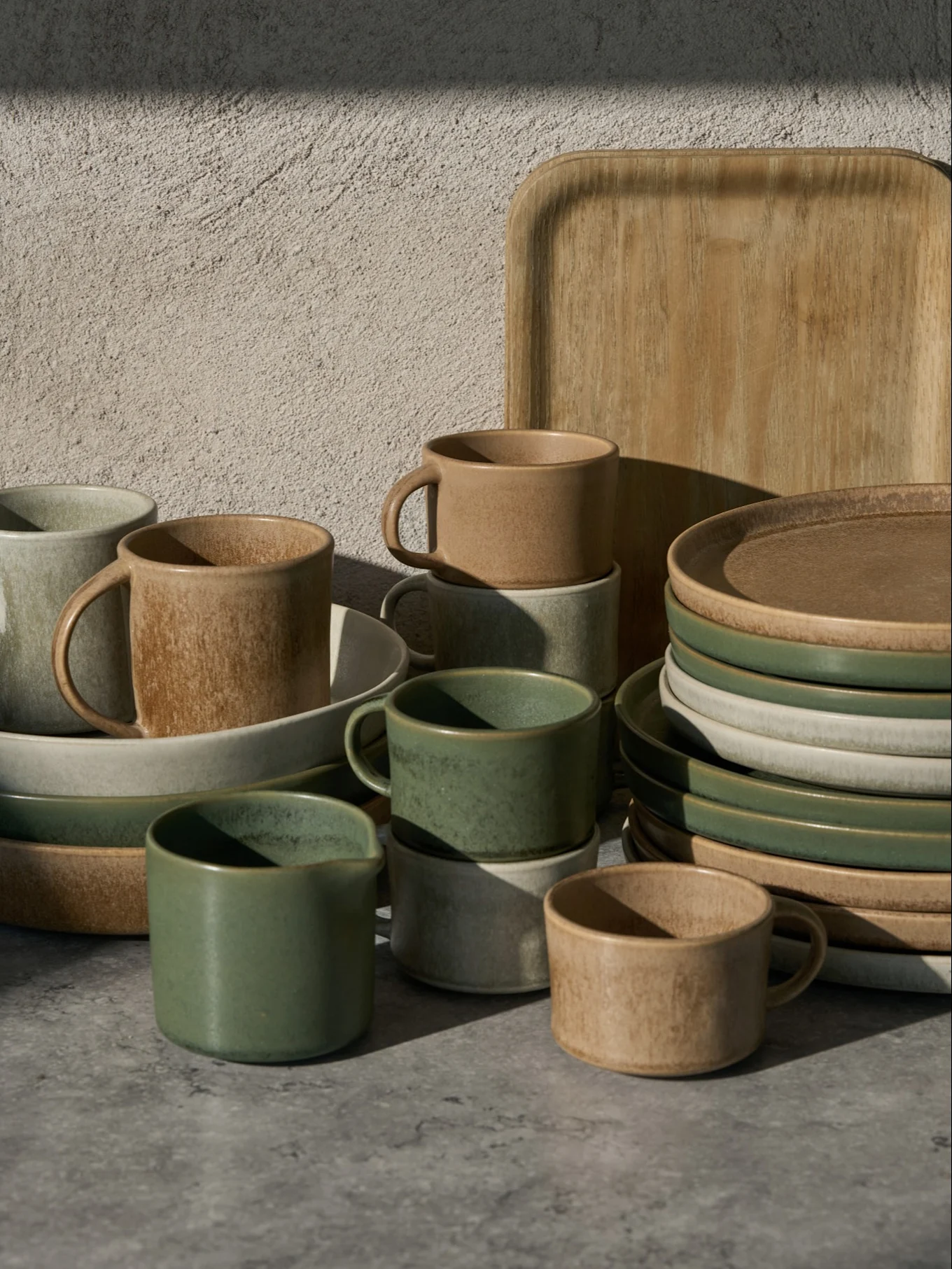 Collectie groene, beige en lichtgrijze keramische kopjes, kommen en borden, met een houten dienblad, op een grijze ondergrond.