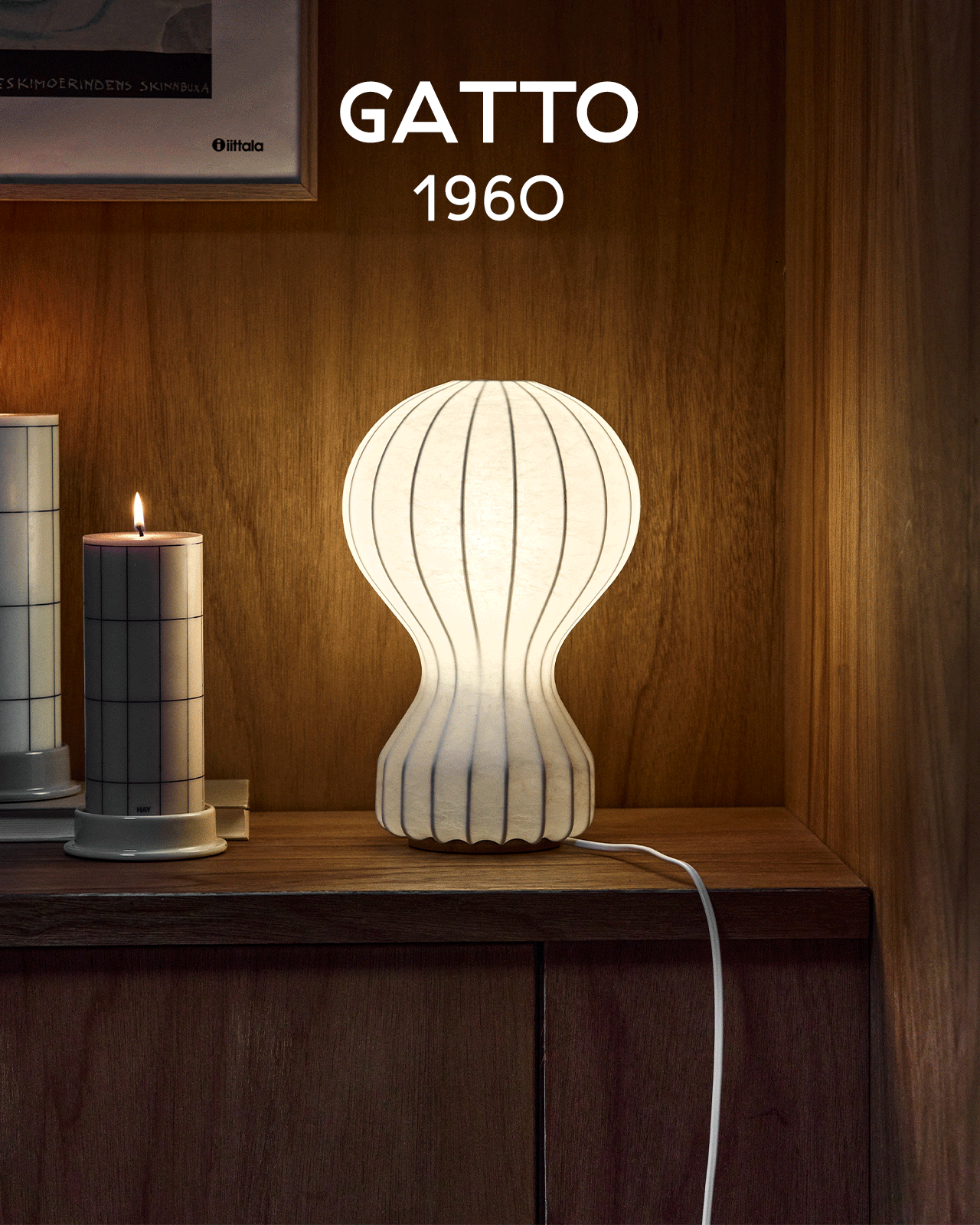 Een gloeiende witte geribbelde lamp in zandlopervorm staat op een houten plank naast een brandende kaars. Tekst: GATTO 1960.