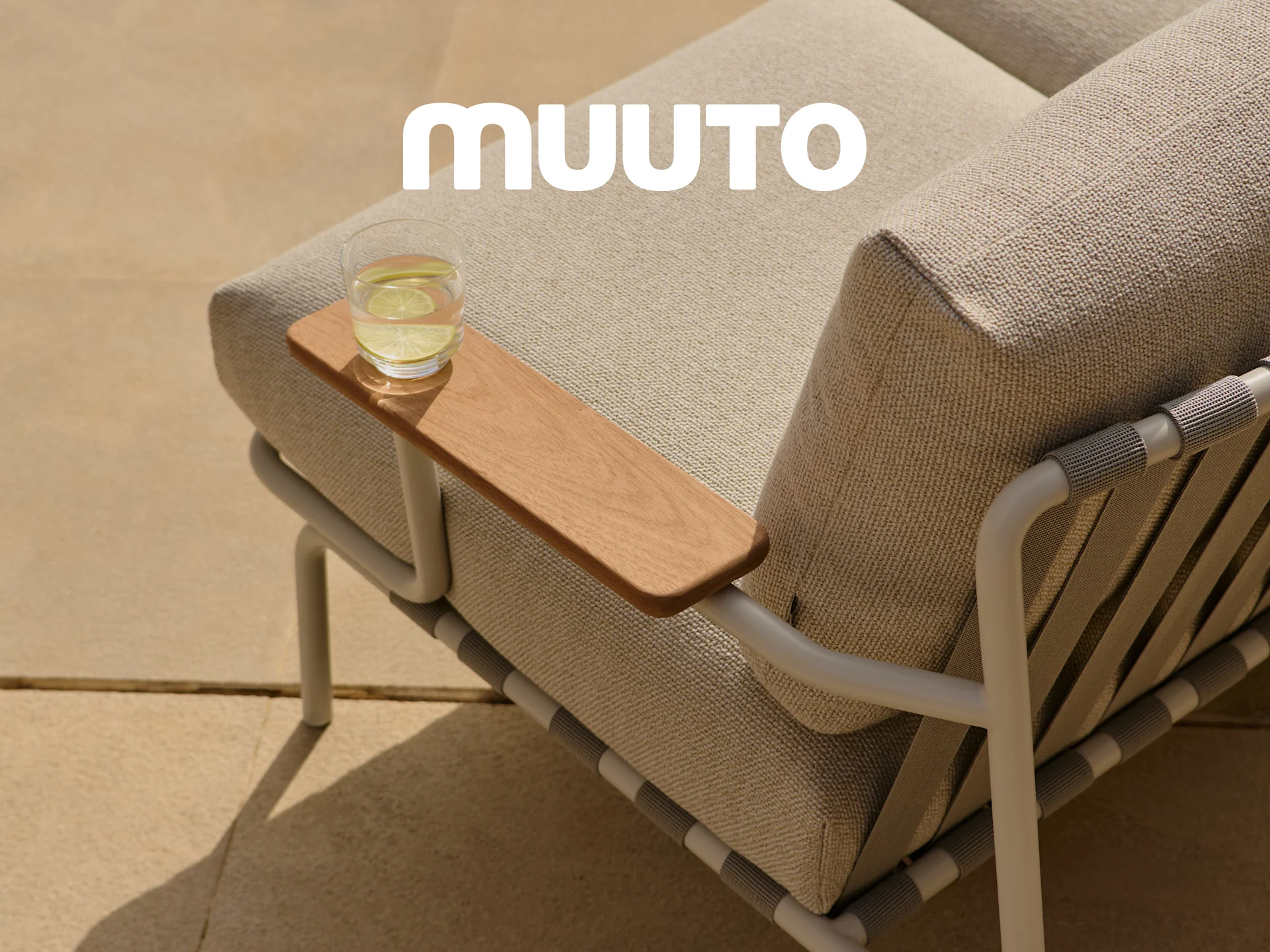 Close-up van een buitenbank met lichte stofkussens, een houten armleuning met een glas water en het witte Muuto-logo.