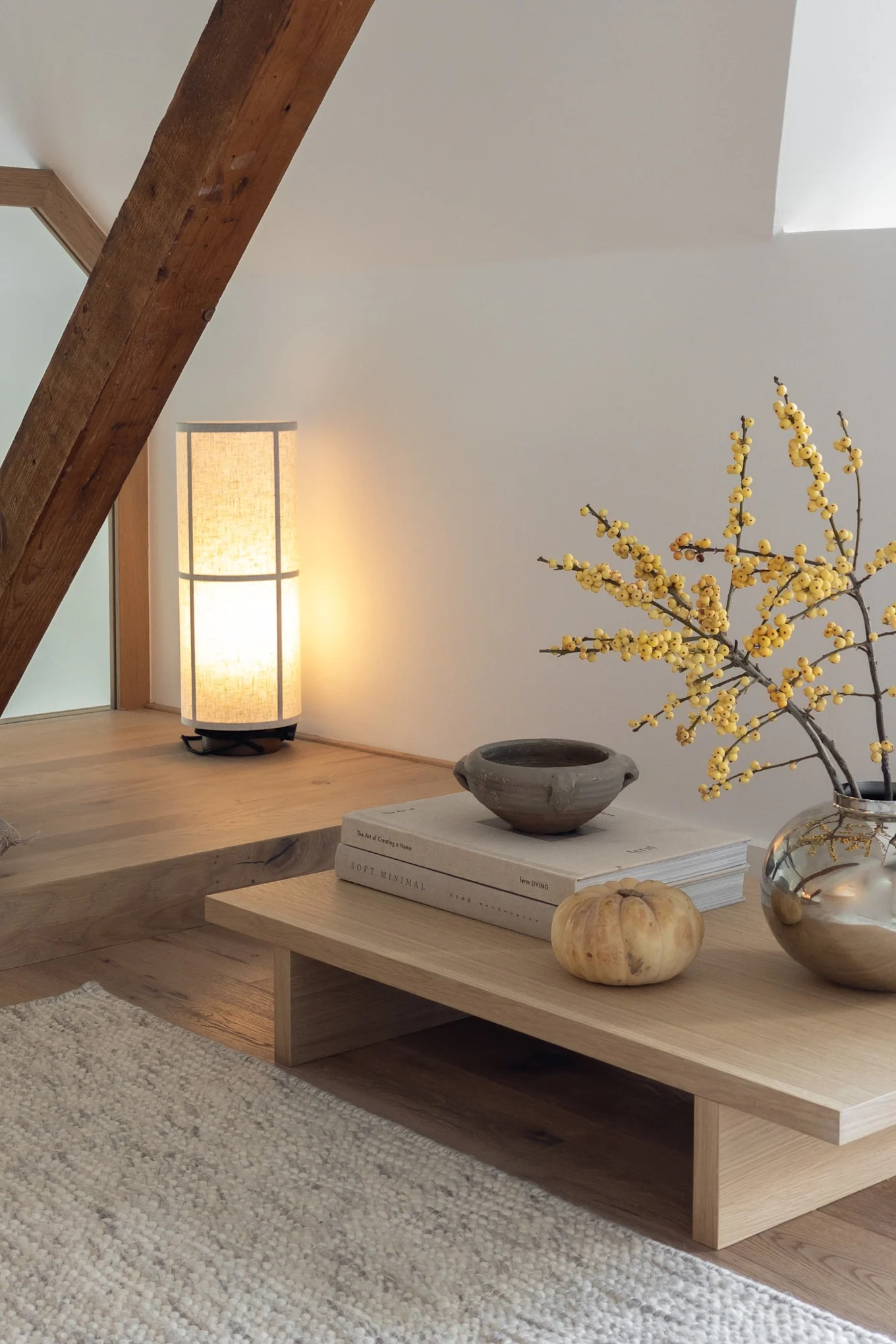 Geometrig zolder woonruimte met hoge lamp, houten balk, lage salontafel met gele bessen, boeken en een pompoen.