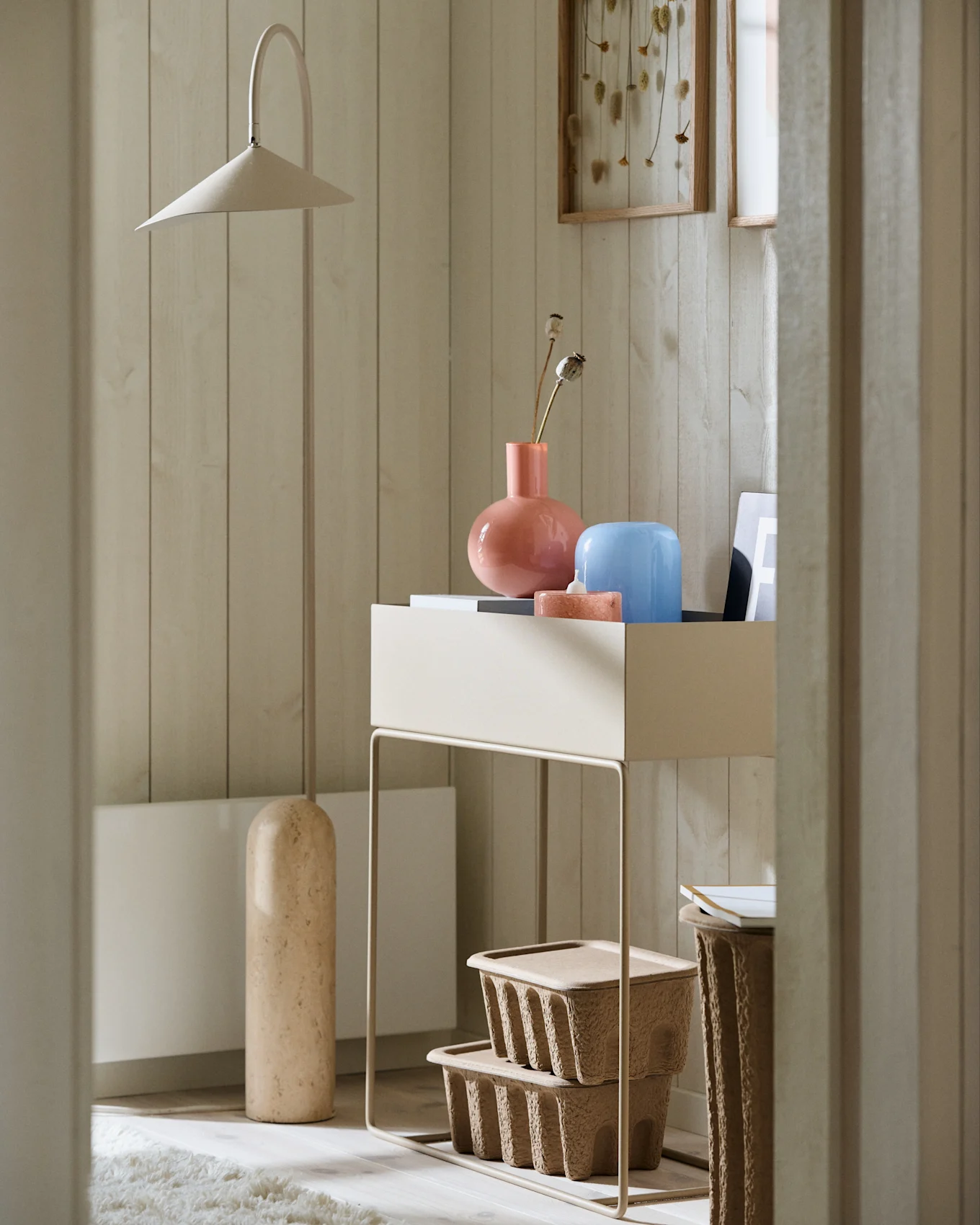 Minimalistische hoek met een hoge beige vloerlamp, een consoletafel met roze en blauwe vazen en getextureerde bruine opbergdozen.
