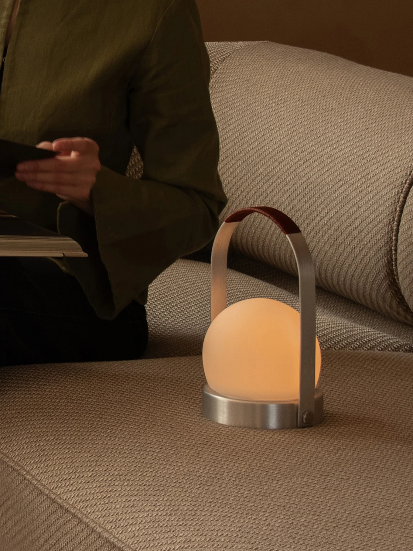 Persoon leest op een bank met een gloeiende draagbare lamp met een matte bol, metalen voet en lederen handvat.