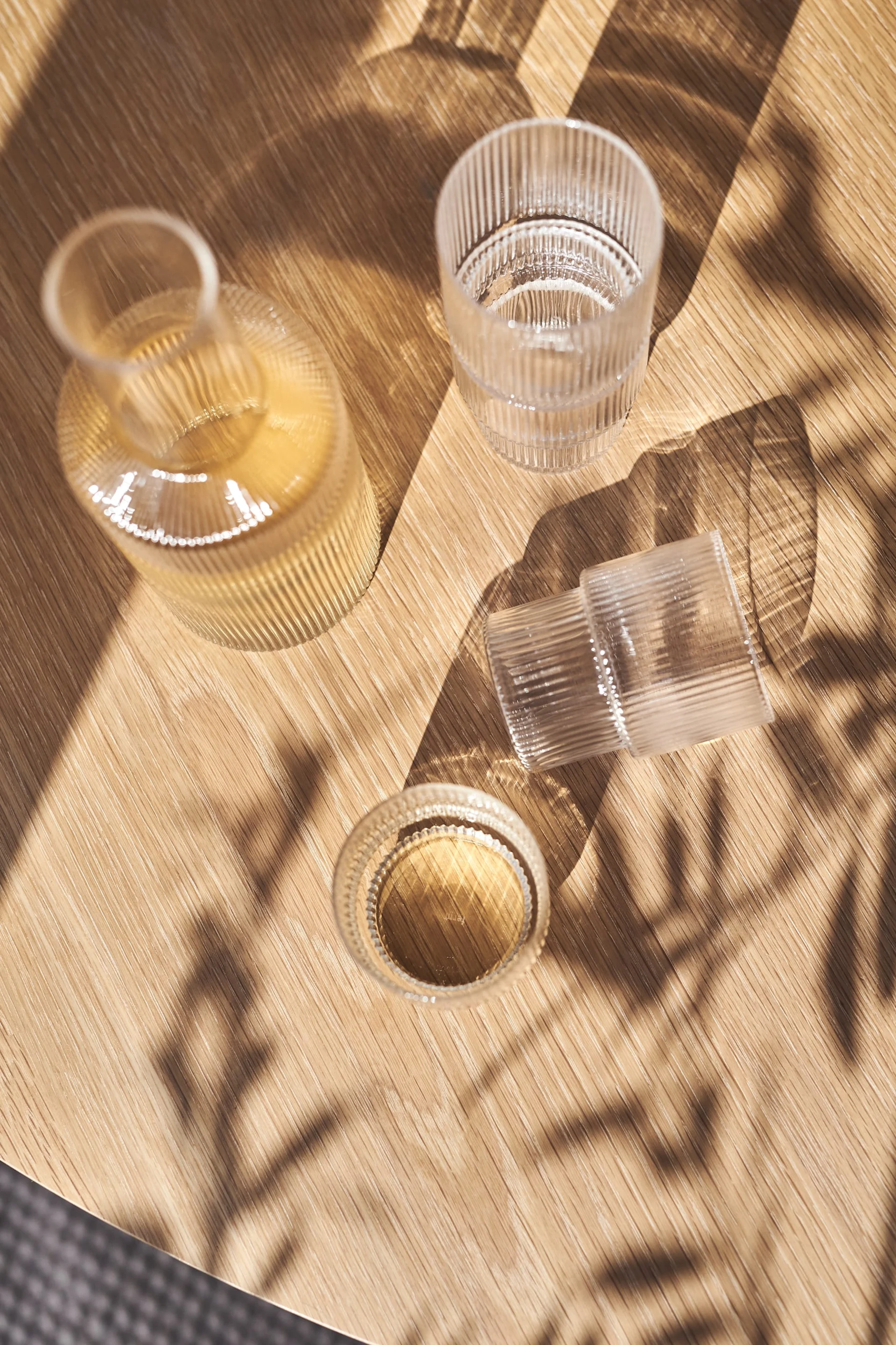 Een geribbelde glazen fles en twee geribbelde glazen, één met vloeistof, op een zonovergoten houten tafel met bladschaduwen.