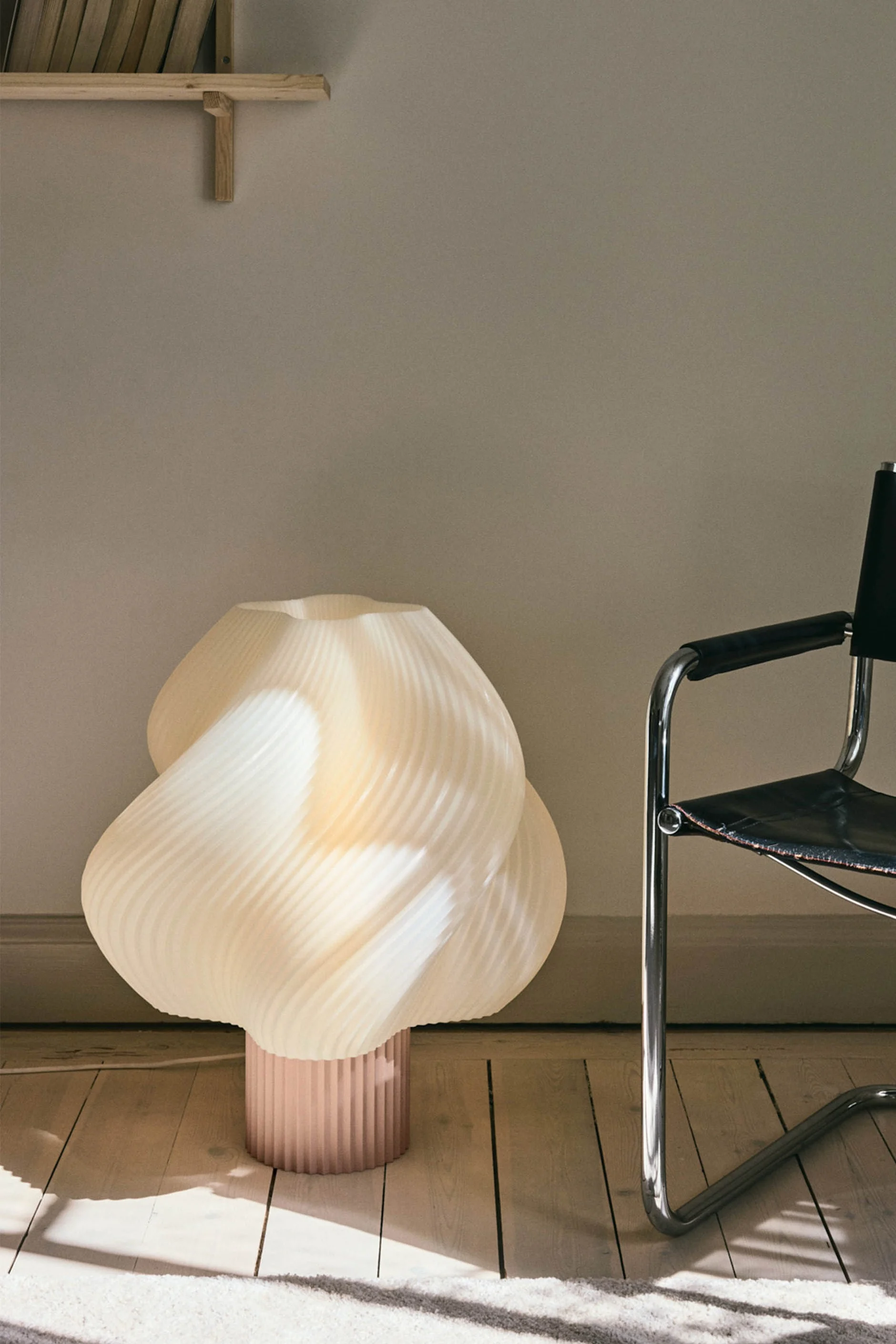 Een sculpturale, geplooide crèmekleurige vloerlamp met een roze basis staat op een houten vloer naast een zwarte leren en chromen stoel.