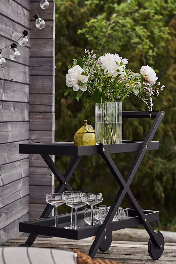 De zwarte Exit serveerwagen van Design House Stockholm staat op een patio en is gevuld met een vaas, bloemen en glazen.