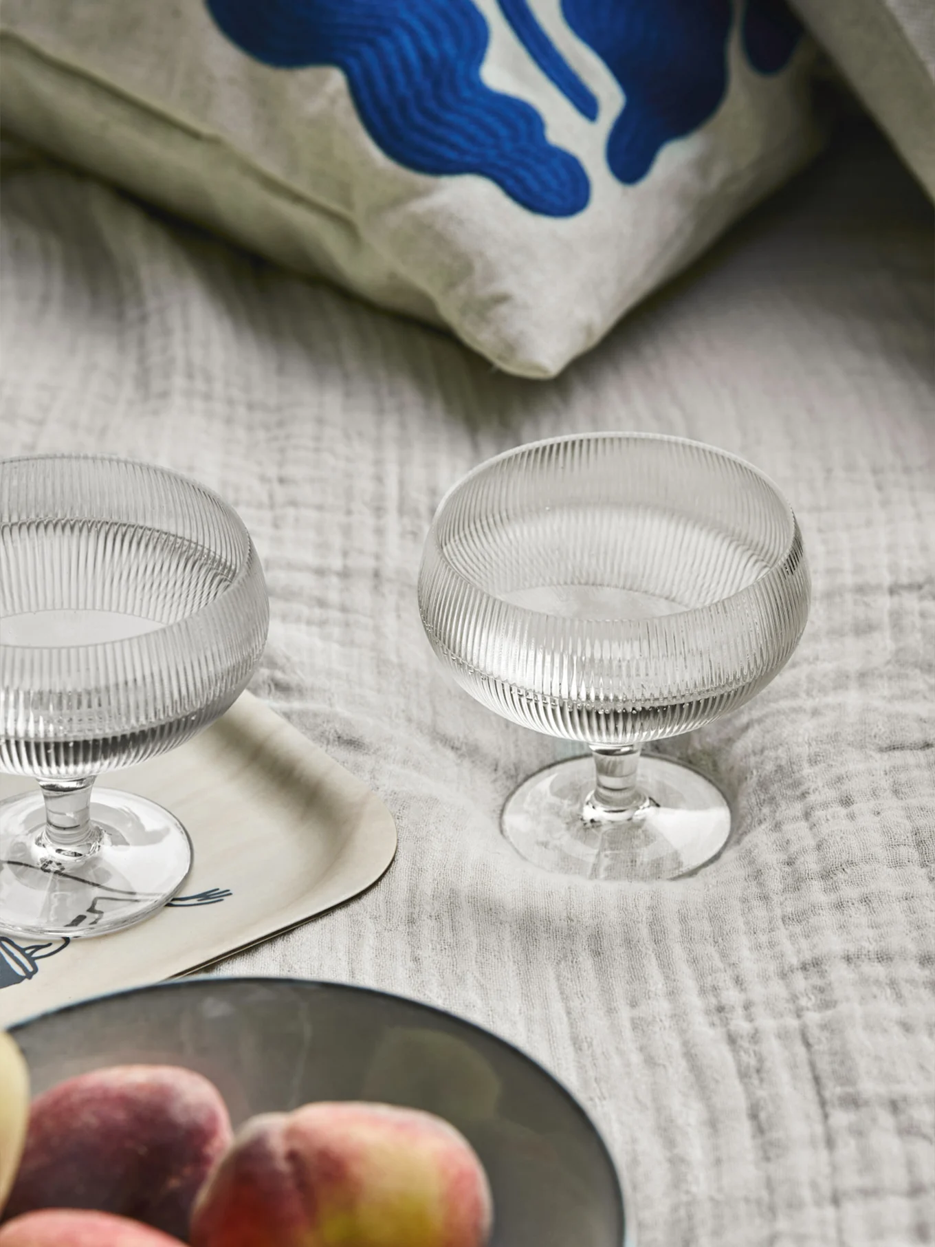 Twee geribbelde glazen dessertkommen, een kussen met blauwe abstracte kunst, een houten dienblad en perziken op een lichte stof.