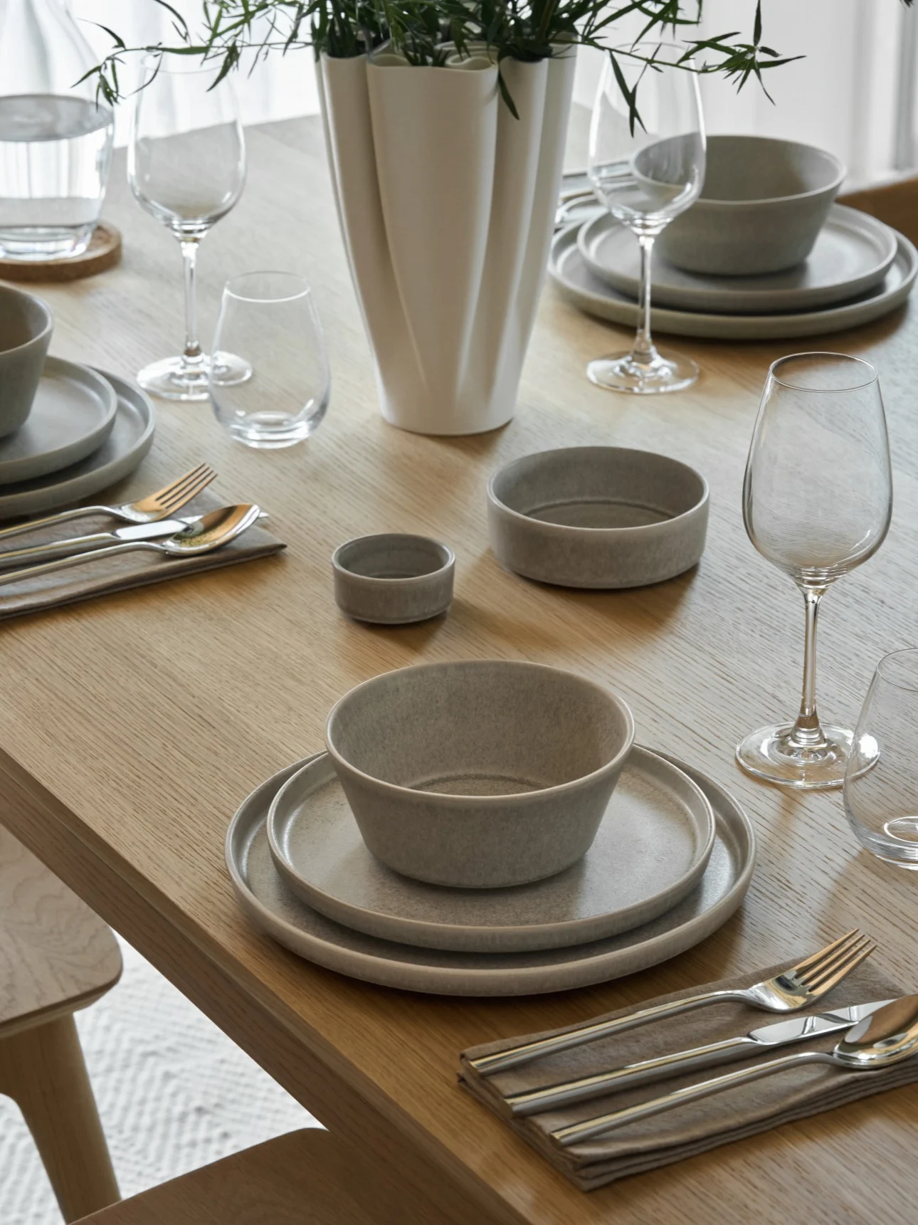 Een lichte houten eettafel gedekt met grijs keramisch servies, zilveren bestek, heldere glazen en een witte vaas met groen loof.