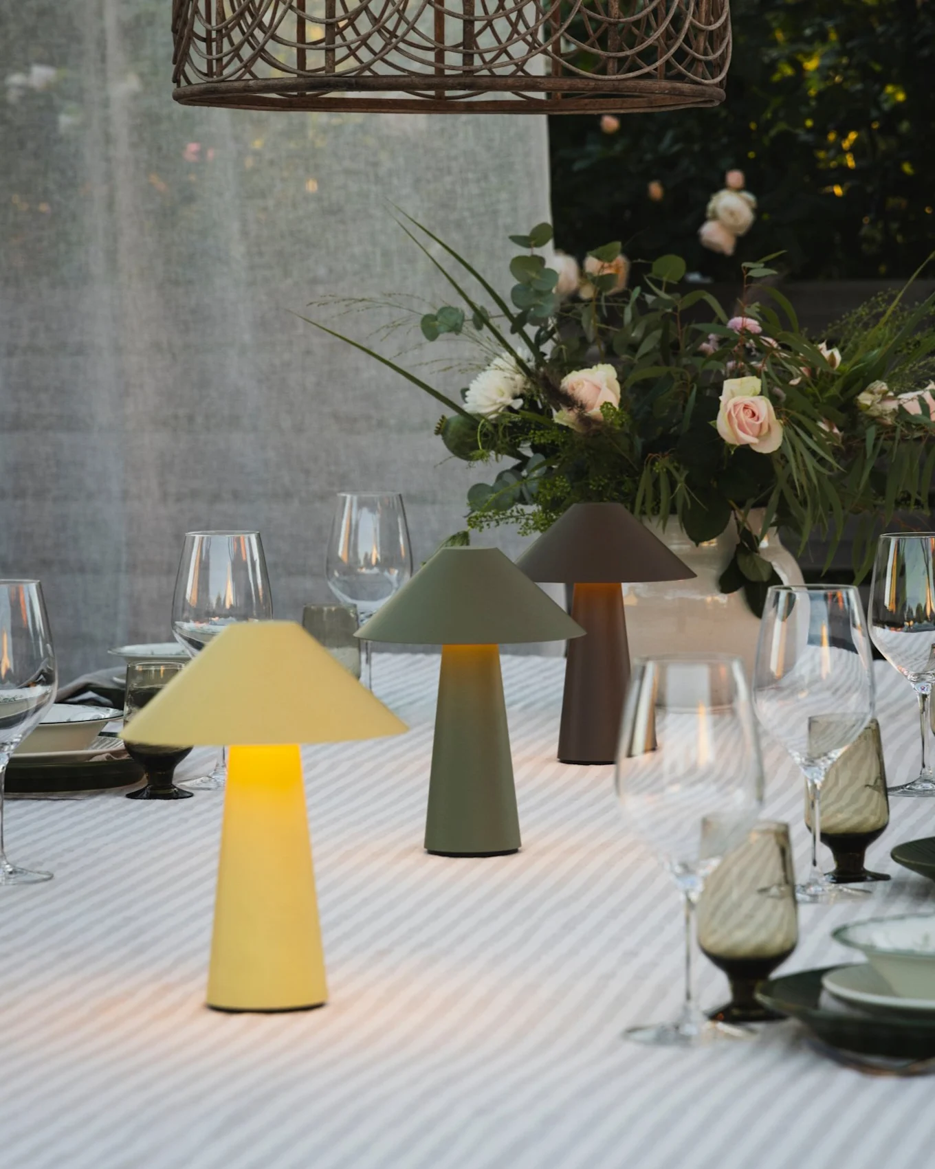 Buiten eettafel met drie verlichte paddenstoellampen, bloemstuk en een rieten hanglamp.