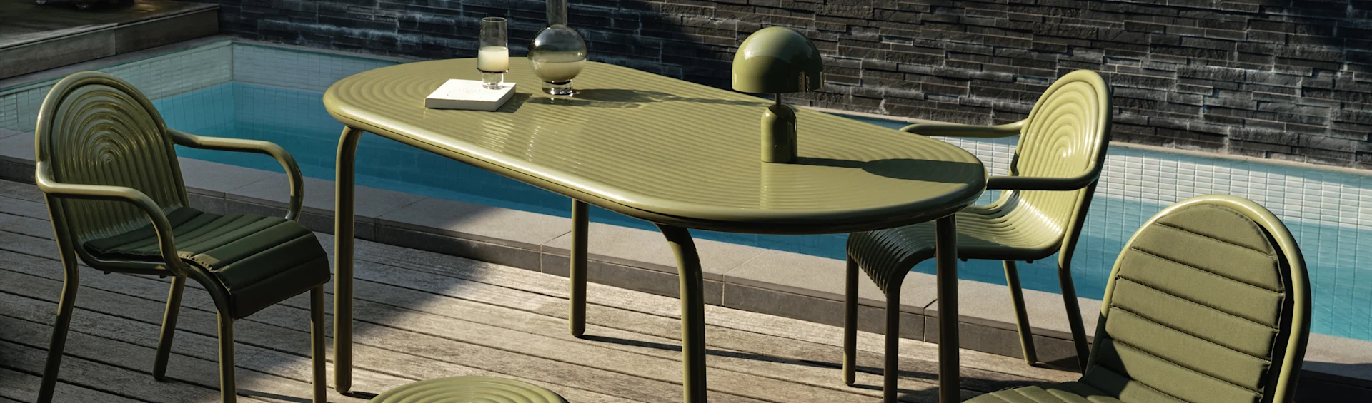 Groene geribbelde buiten eettafel en stoelen op een houten terras bij een zwembad. Een lamp, karaf, glas en boek liggen op tafel.