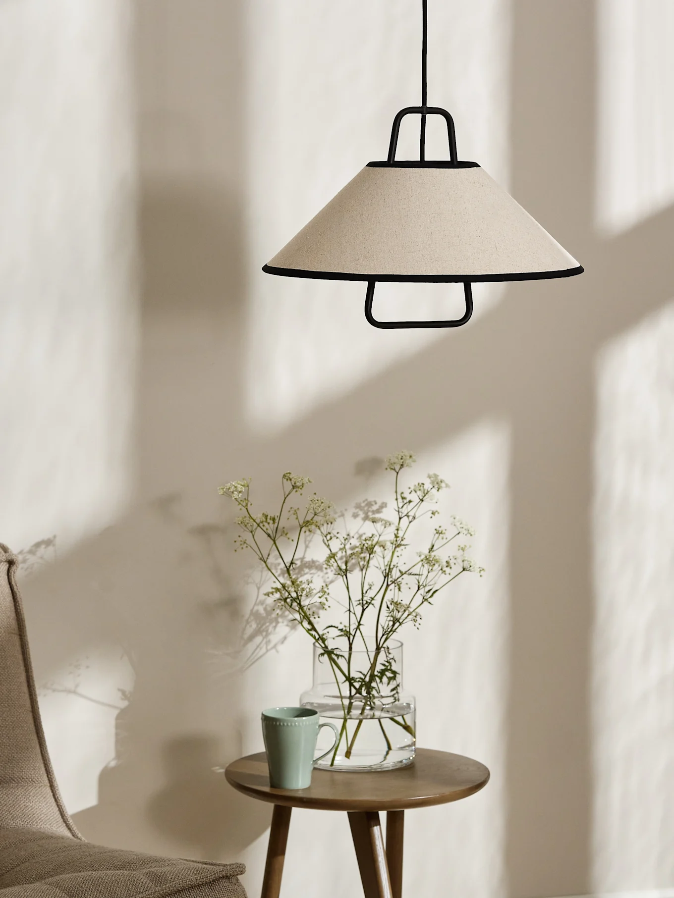 Een conische crèmekleurige hanglamp met zwarte rand hangt boven een houten bijzettafel met een vaas witte bloemen en een lichtgroene mok.