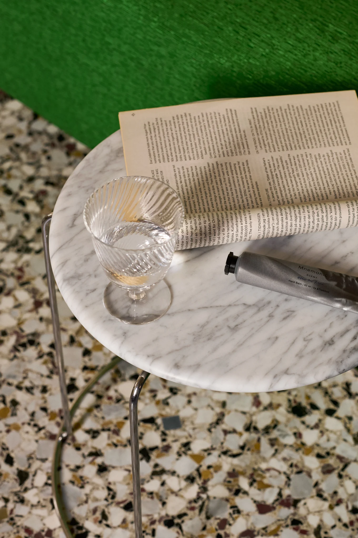 Een helder, geribbeld glas water, een open boek en een zilveren handbalsemtube op een marmeren tafel.
