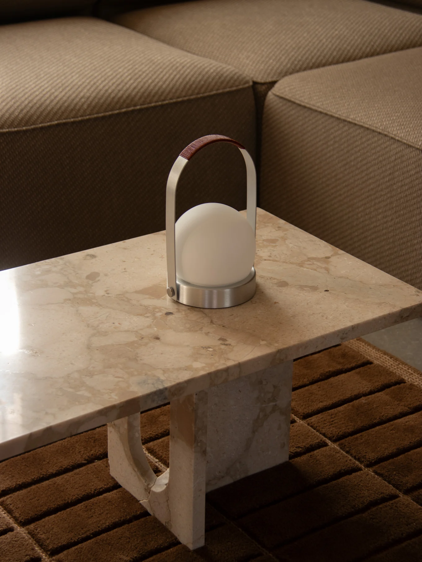 Moderne draagbare lamp met een bolvormig wit licht, zilveren frame en bruin leren handvat op een marmeren tafel naast een bank.