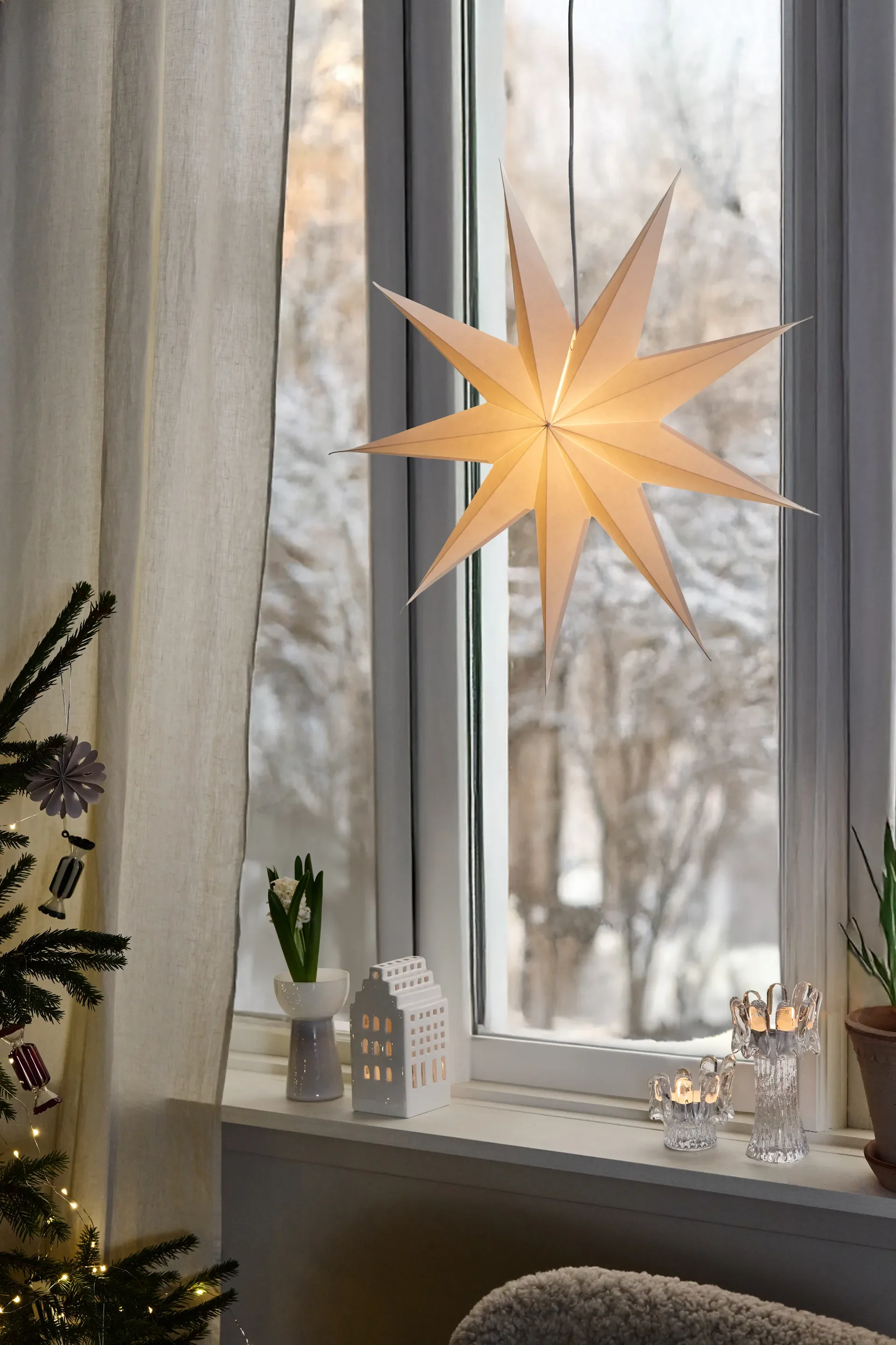 Winterversierd raam met gloeiende papieren ster, keramisch huis, brandende kaarsen en kerstboom.