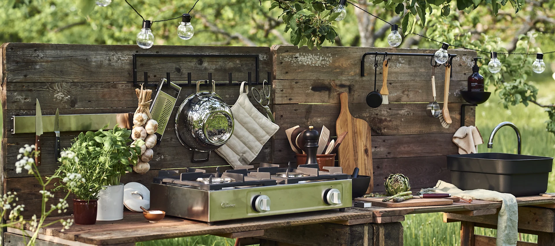 Buitenkeuken van rustieke houten planken met een groen draagbaar gasfornuis, kookgerei, kruiden, knoflook en groenten.