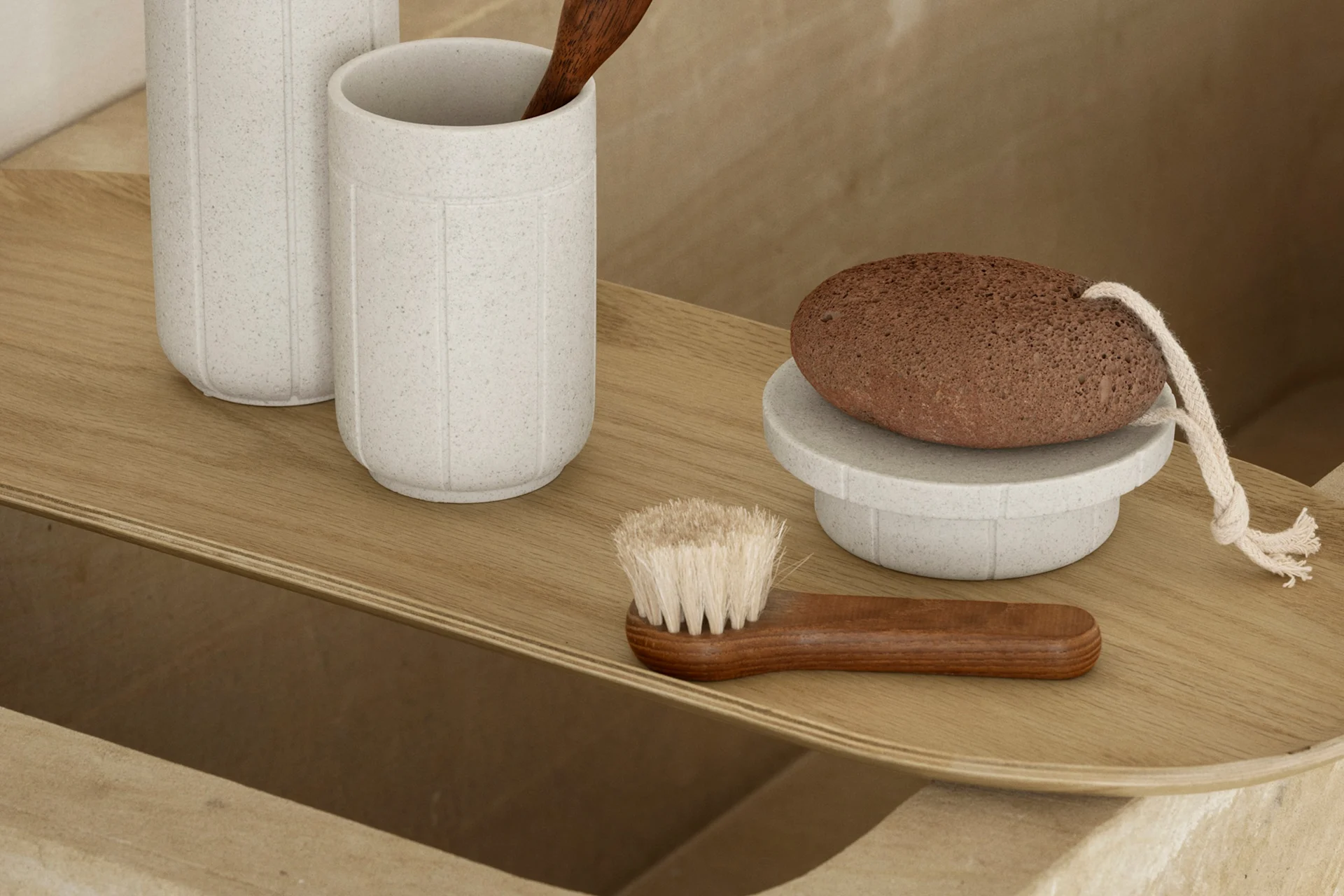 Een licht houten dienblad met badaccessoires: twee gespikkelde grijze bekers, een houten lepel, een bruine puimsteen op een standaard en een houten borstel.