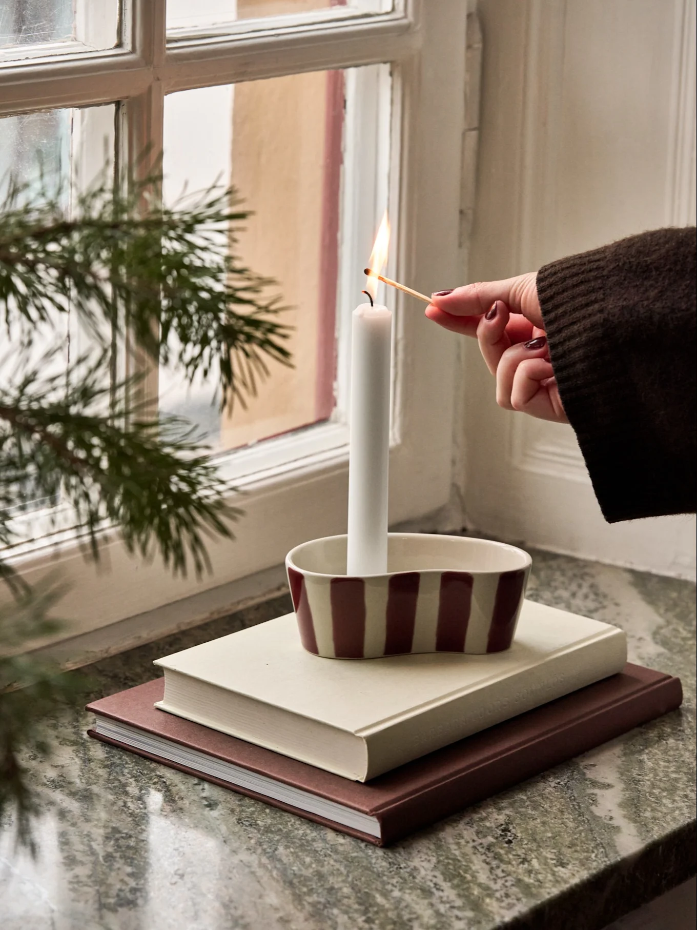 Een hand steekt een witte kaars aan in een gestreepte houder op een stapel boeken bij een raam met dennentakken.