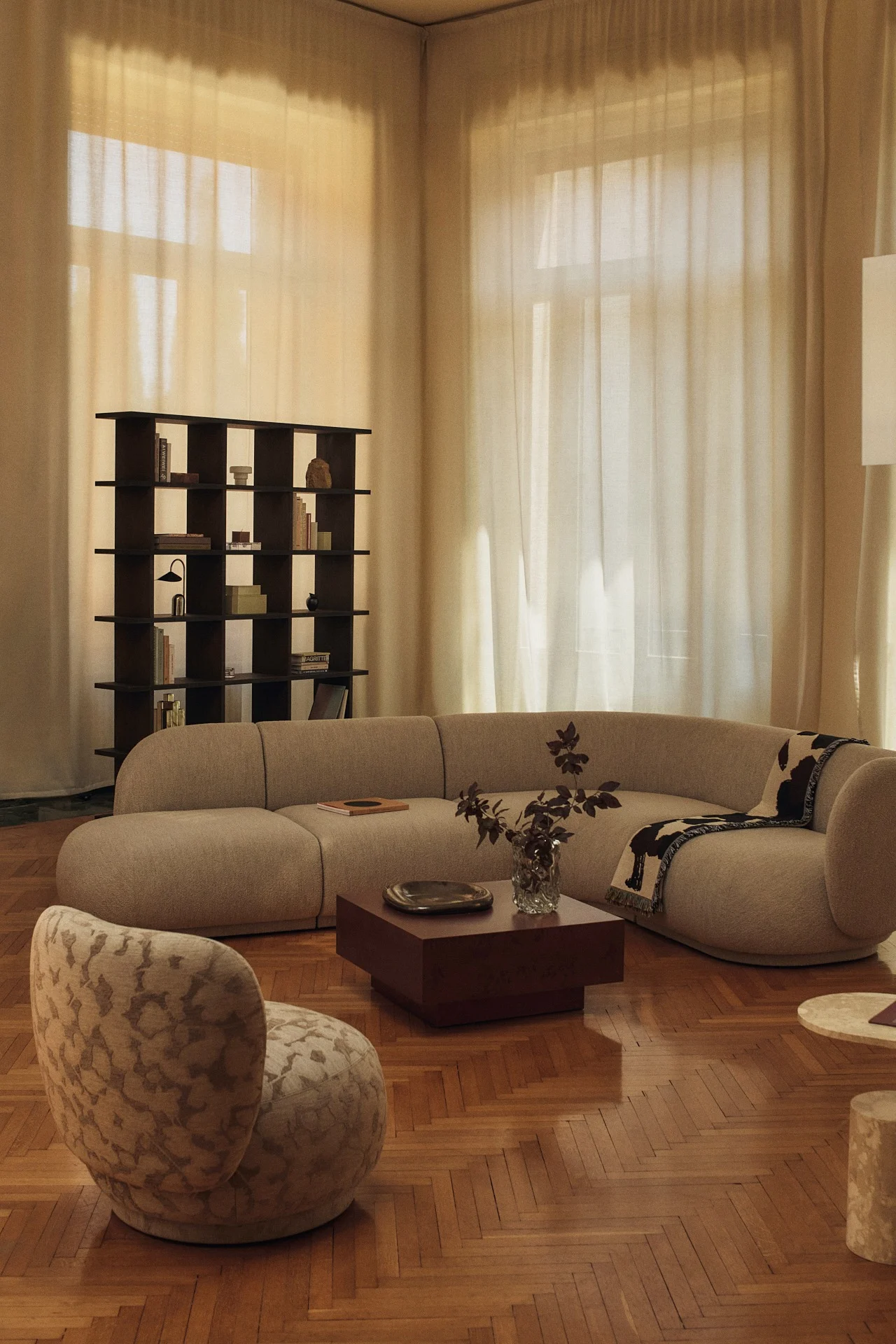 Een woonkamer met een gebogen beige modulaire bank, een donkere vierkante salontafel, een donkere open boekenkast en een gestoffeerde accentstoel op een parketvloer.