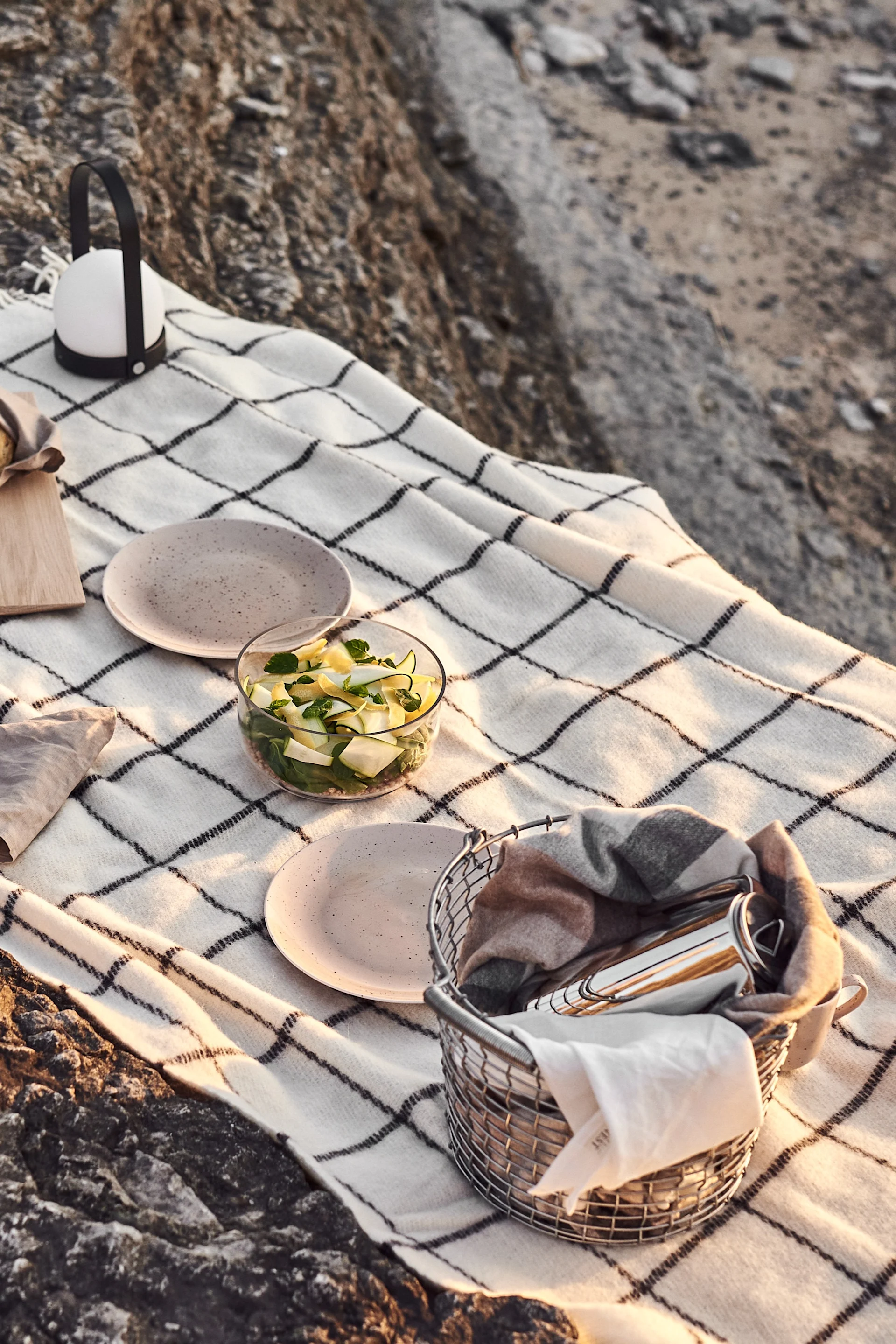 Picknickkleed op rotsen met salade, borden, een lantaarn en een mand met bestek.