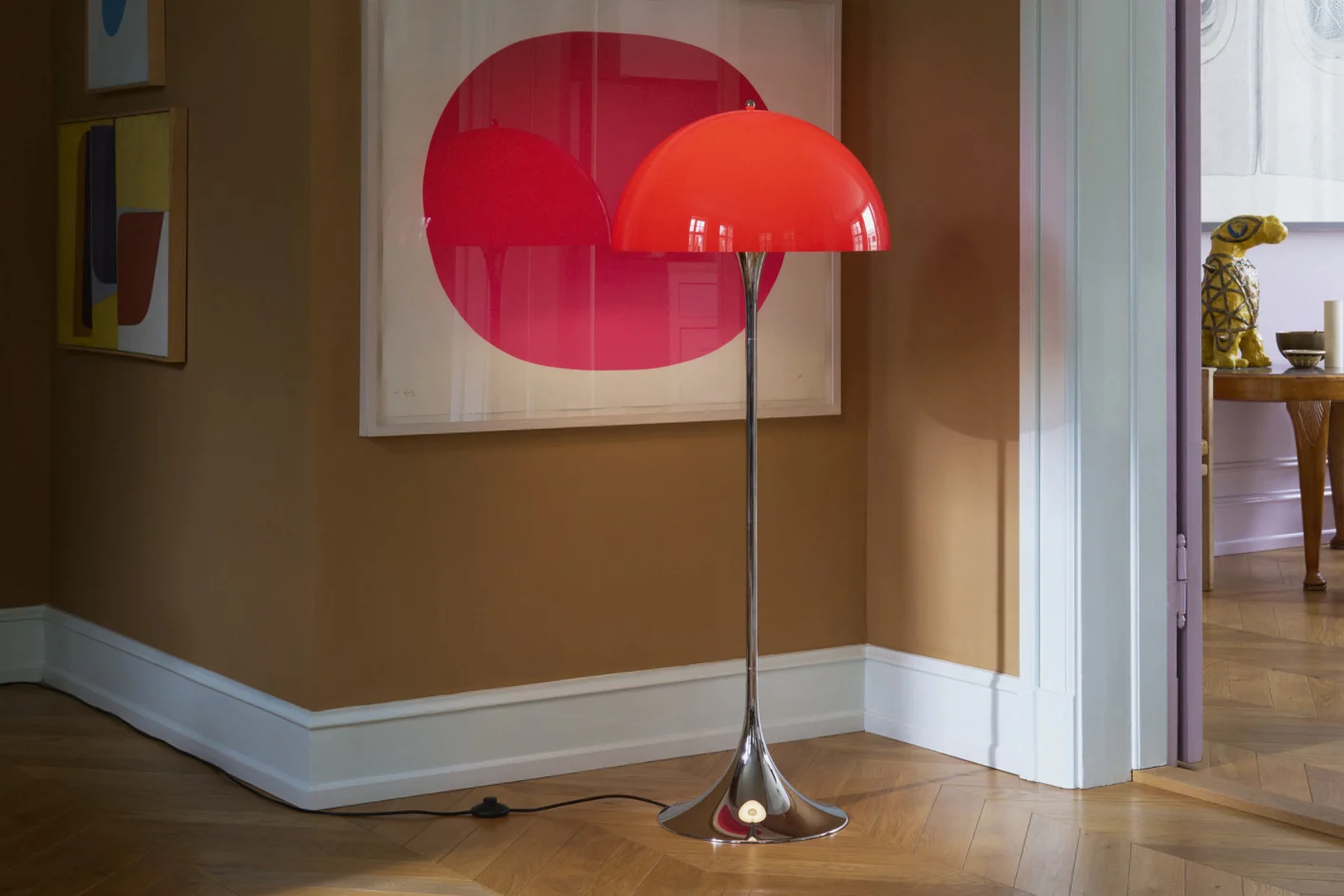 Een felrode koepelvormige vloerlamp met chromen standaard staat op een houten vloer in een kamer met bruine muren en moderne kunst.