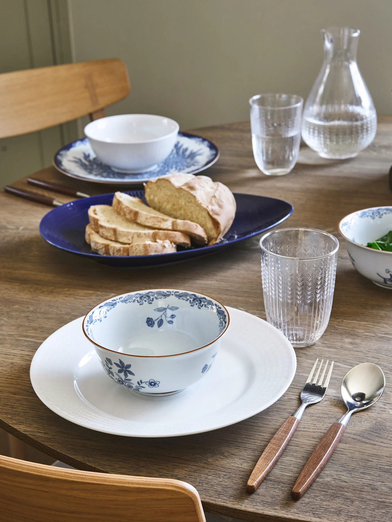 Eettafel gedekt met twee couverts, blauw-witte bloemschalen, brood op een blauw bord en bestek met houten handvatten.