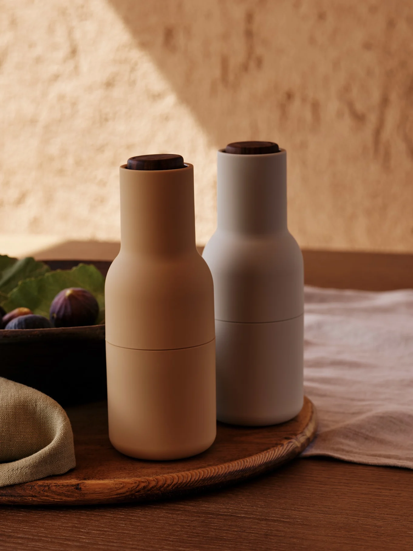Twee moderne kruidenmolens met houten doppen, één beige en één lichtgrijs, op een ronde houten schaal met vijgen.
