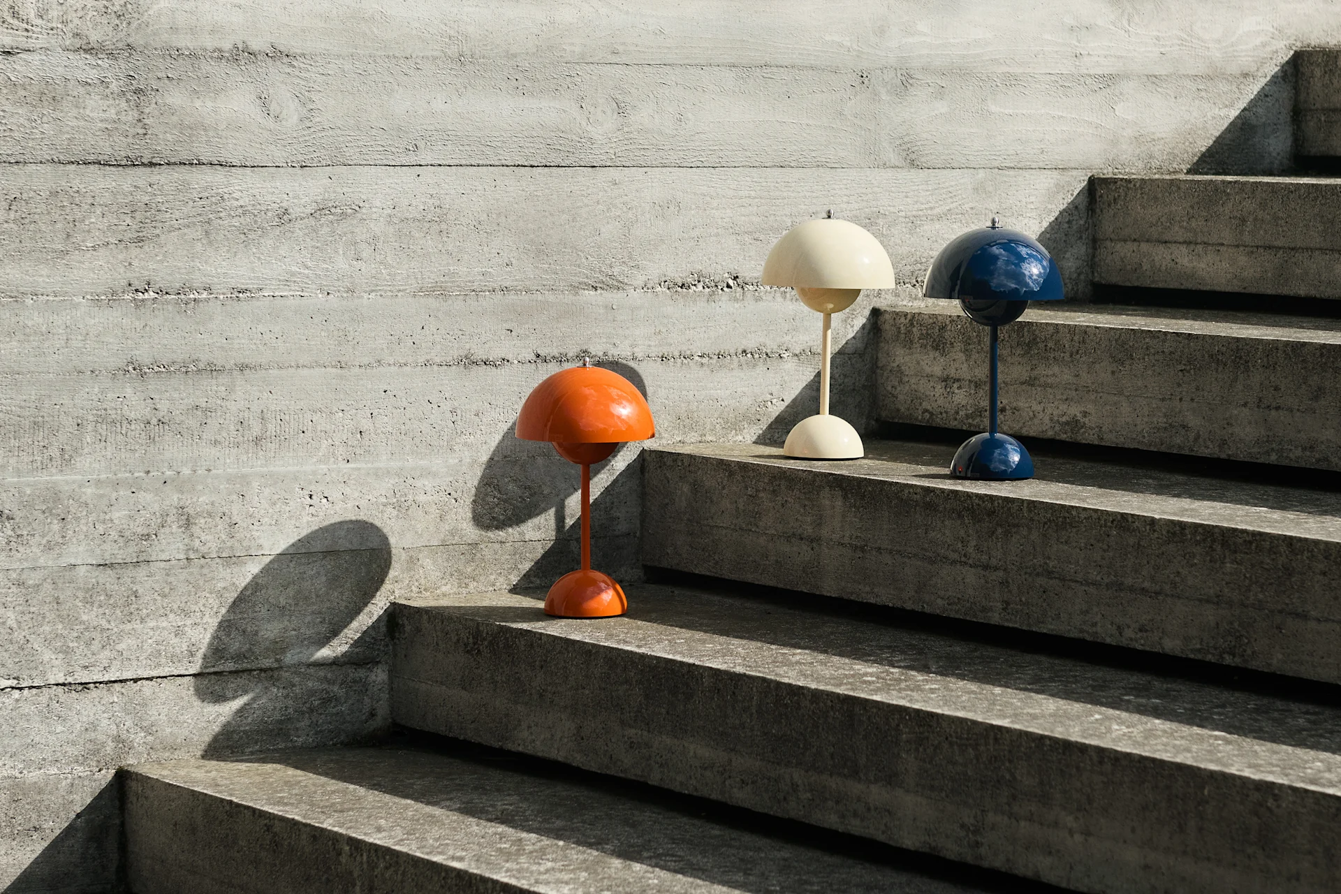 Drie koepelvormige tafellampen in oranje, crème en blauw op betonnen trappen naast een betonnen muur, die schaduwen werpen.