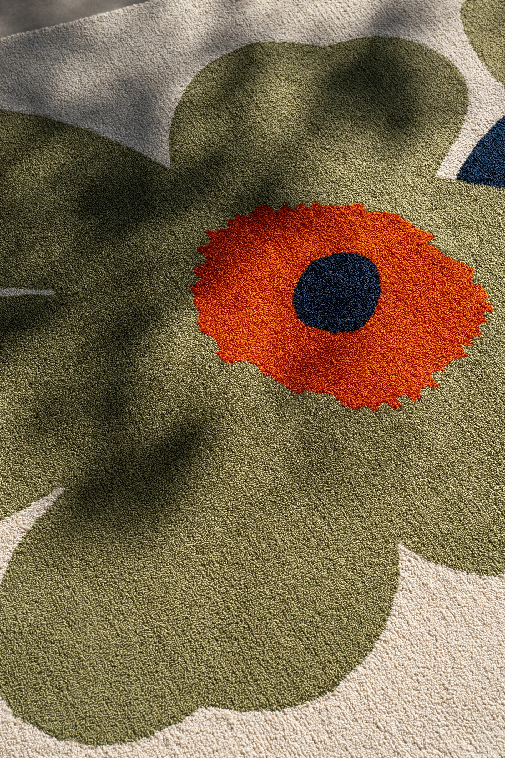 Close-up van een tapijt met een groot, gestileerd bloemendesign. Olijfgroene bloembladen, een oranje hart en een donkerblauwe stip op een crèmekleurige achtergrond.