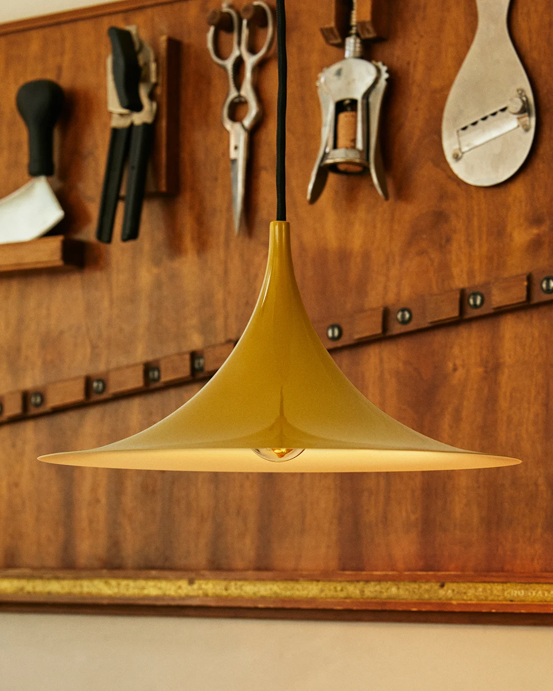 Gouden klokvormige hanglamp met zichtbare gloeilamp, tegen een houten wand met hangende gereedschappen.