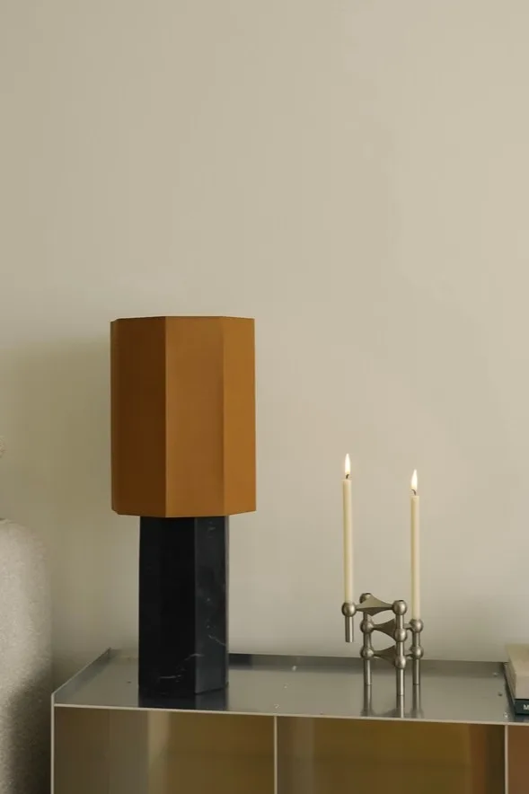 Moderne tafellamp met zeshoekige bruine kap en zwarte marmeren voet, naast een zilveren modulaire kandelaar met twee brandende witte kaarsen.