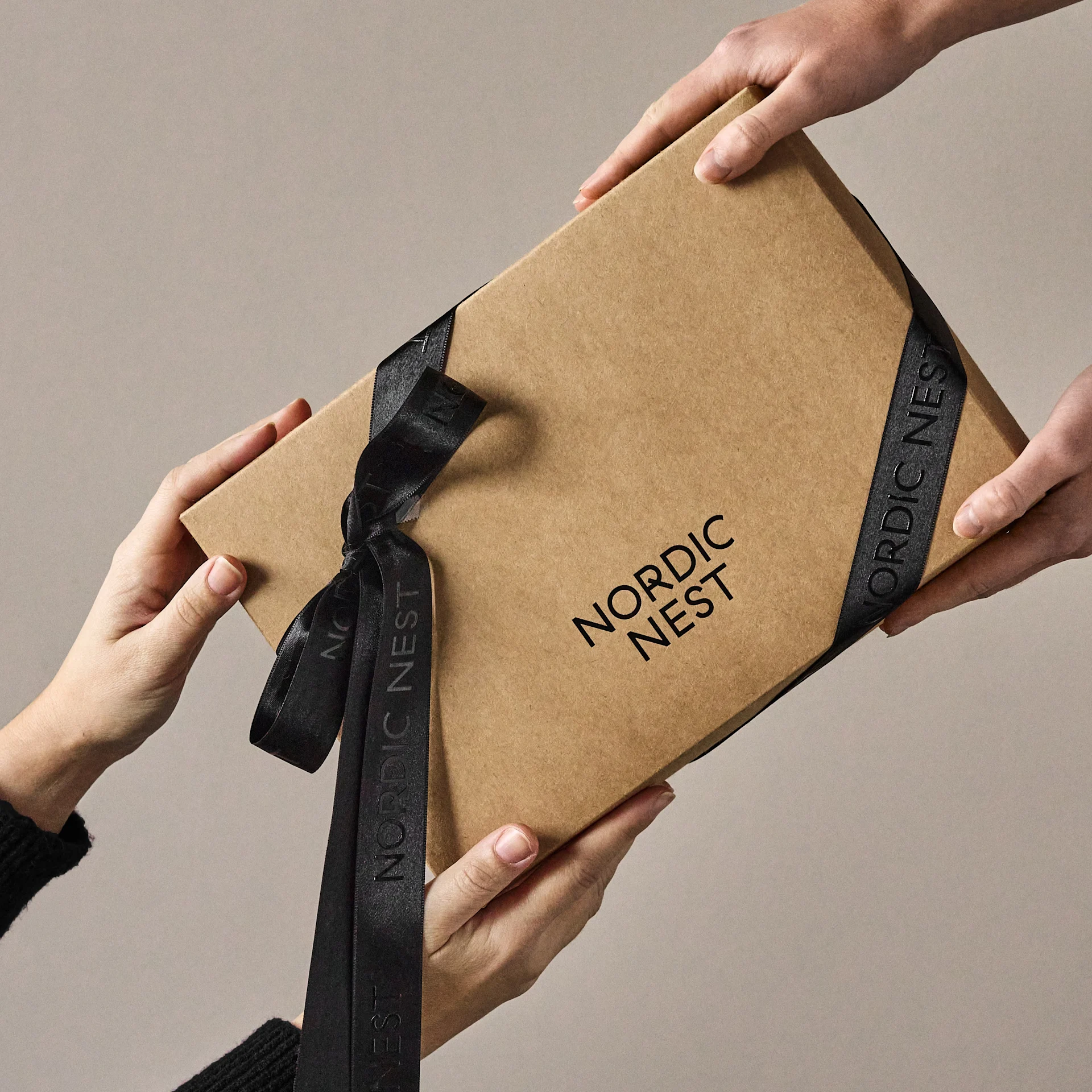 Handen wisselen een bruine geschenkdoos uit met 'Nordic Nest'-logo en zwart lint.