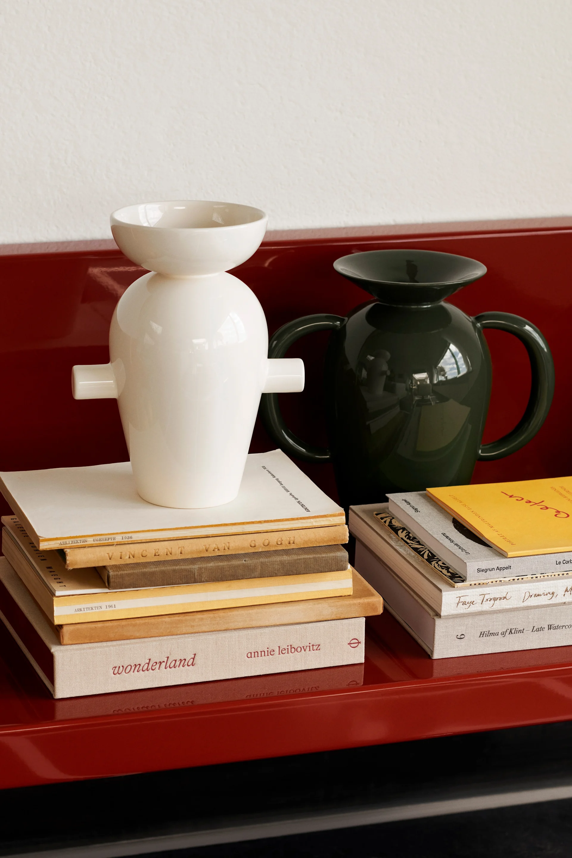 Twee glanzende keramische vazen, één wit en één donkergroen, op stapels boeken op een rood oppervlak.