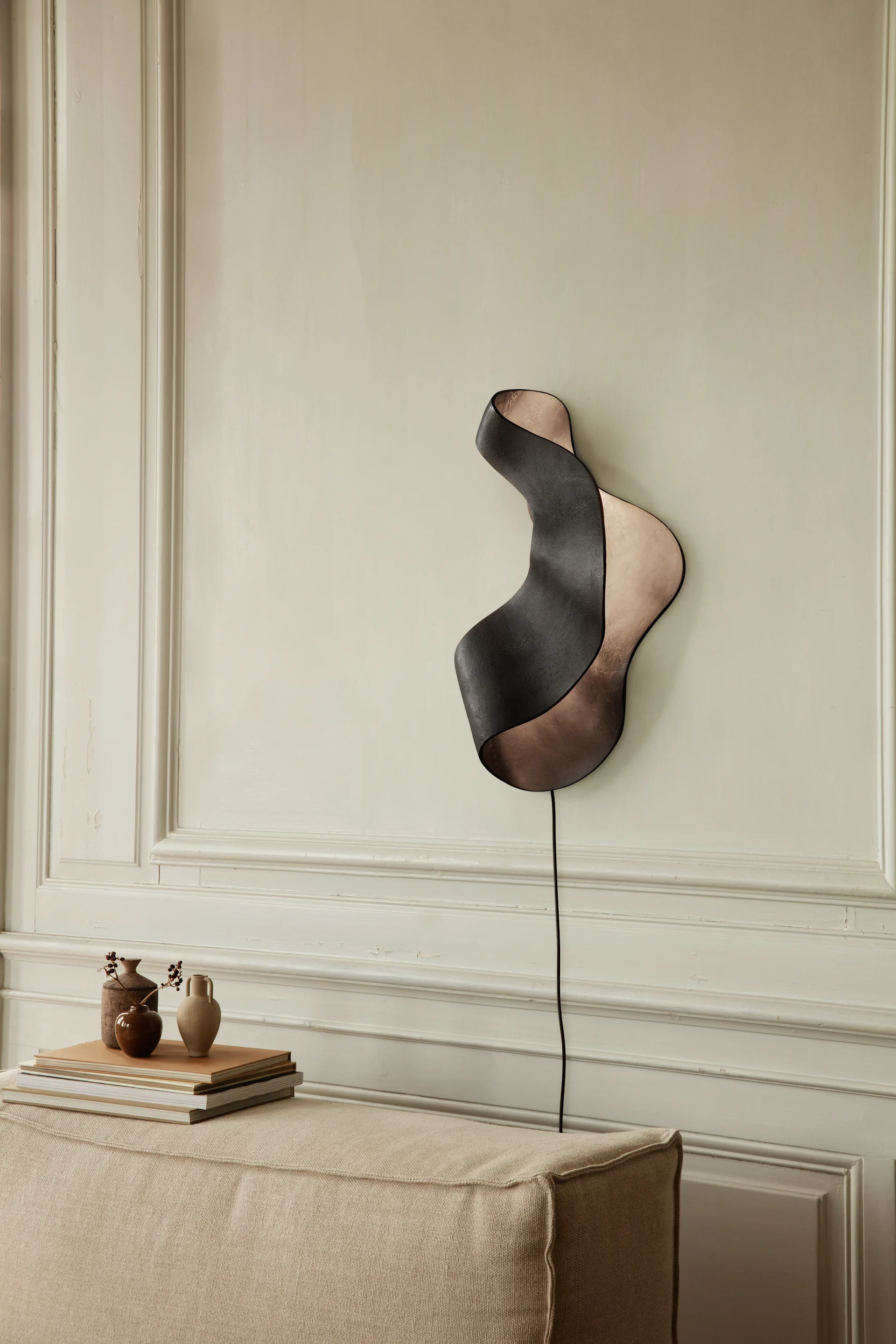 Golvende wandlamp met donkere buitenkant en koperen binnenkant, gemonteerd op een lambrisering boven een stoffen bank.