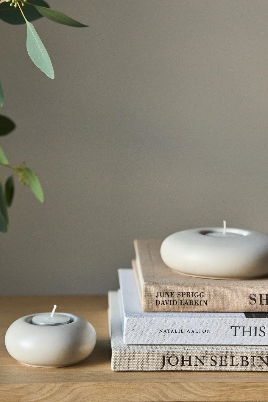 Twee ronde, lichtgekleurde keramische kaarsenhouders, één op een stapel boeken, en een eucalyptus tak.