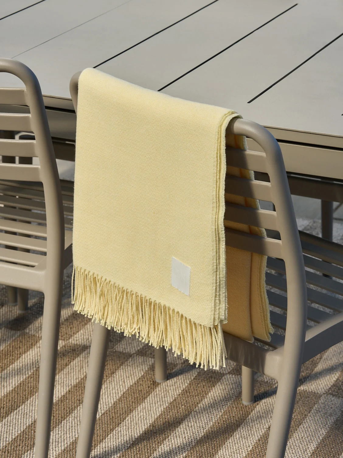 Lichtgele deken met franjes gedrapeerd over een beige buitenstoel.