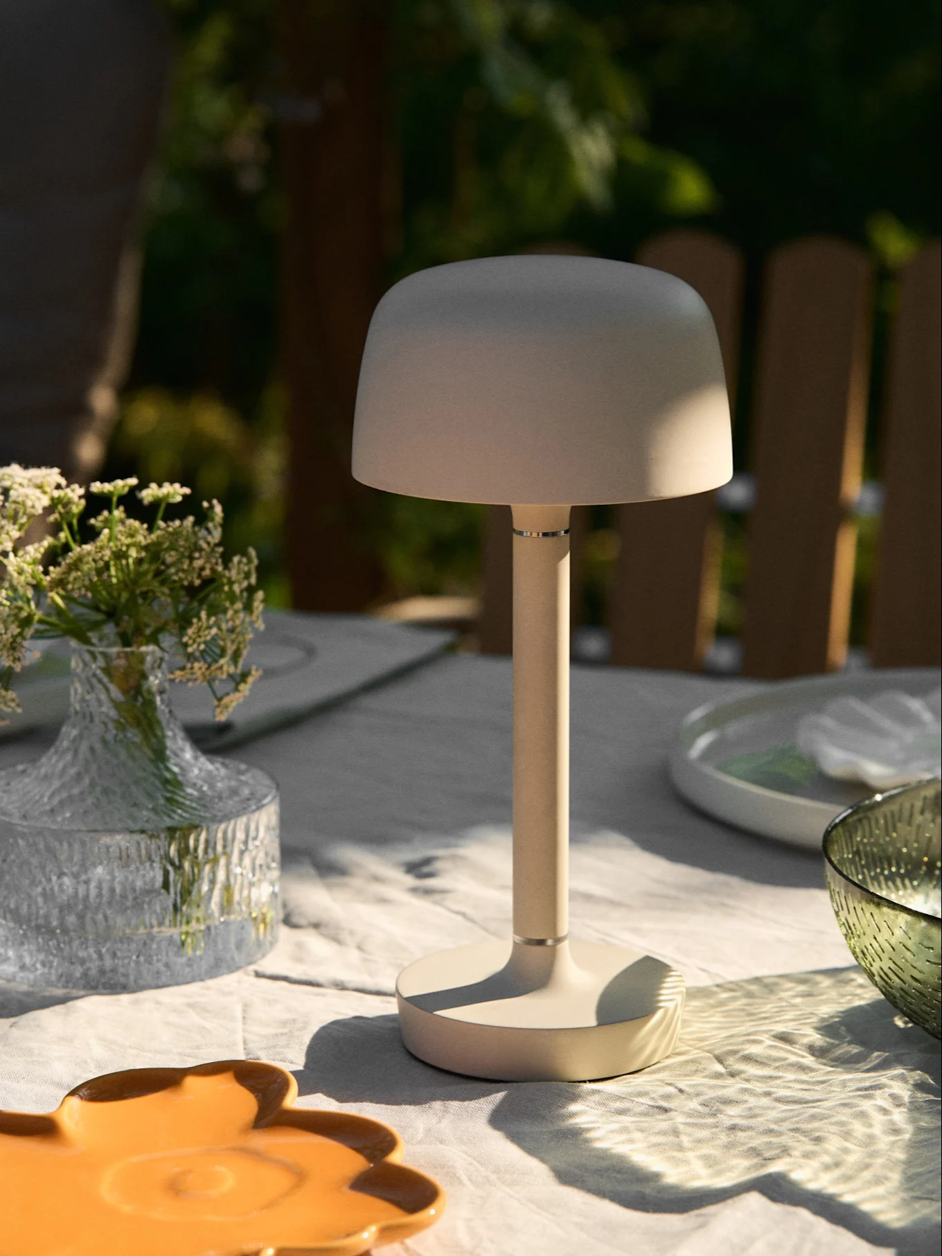 Beige draagbare tafellamp op een buitentafel met bloemenvaas en gevlekt zonlicht.