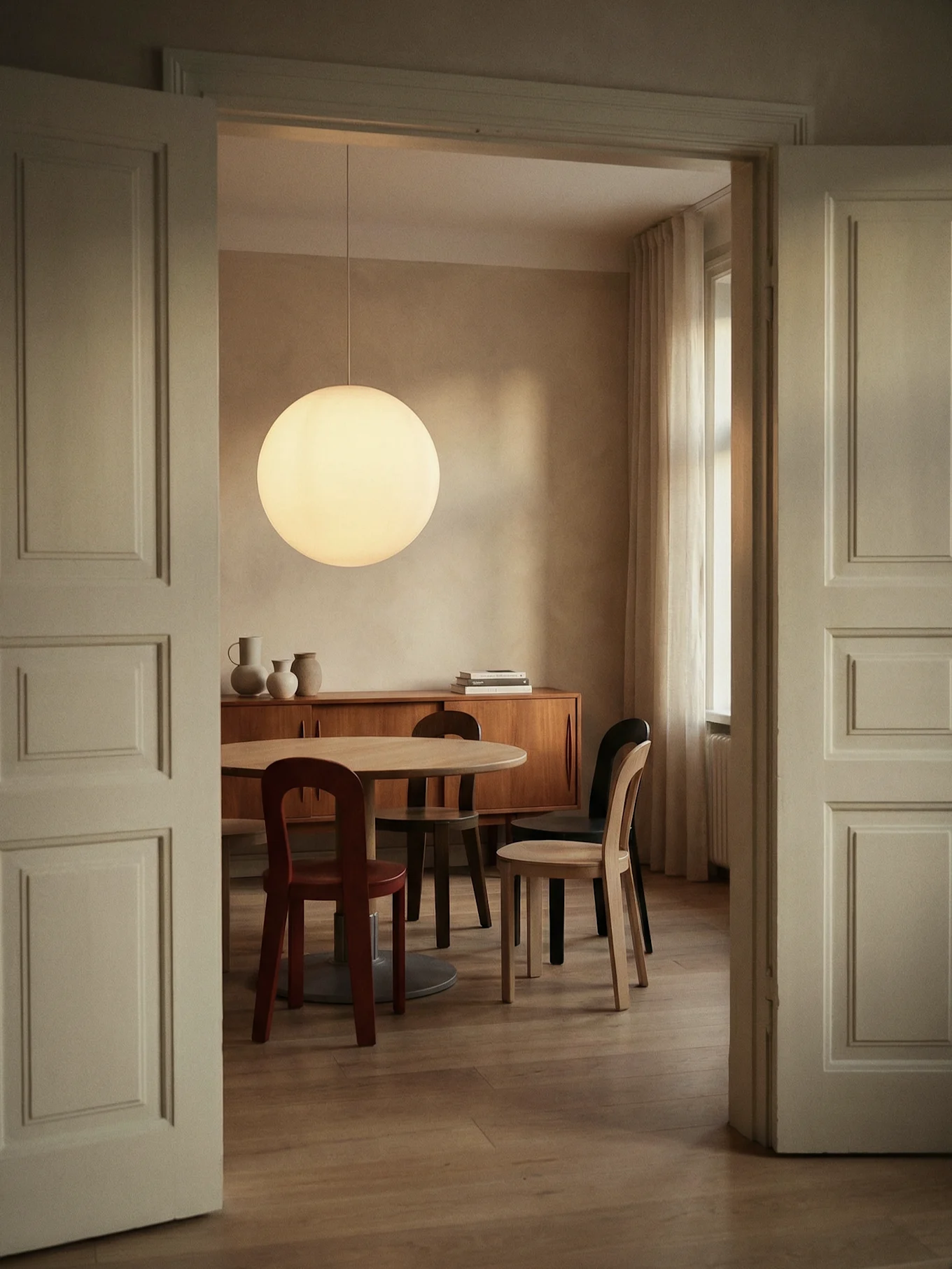 Eetkamer door open deuropening, met ronde houten tafel, diverse stoelen, grote bolvormige hanglamp en houten dressoir.