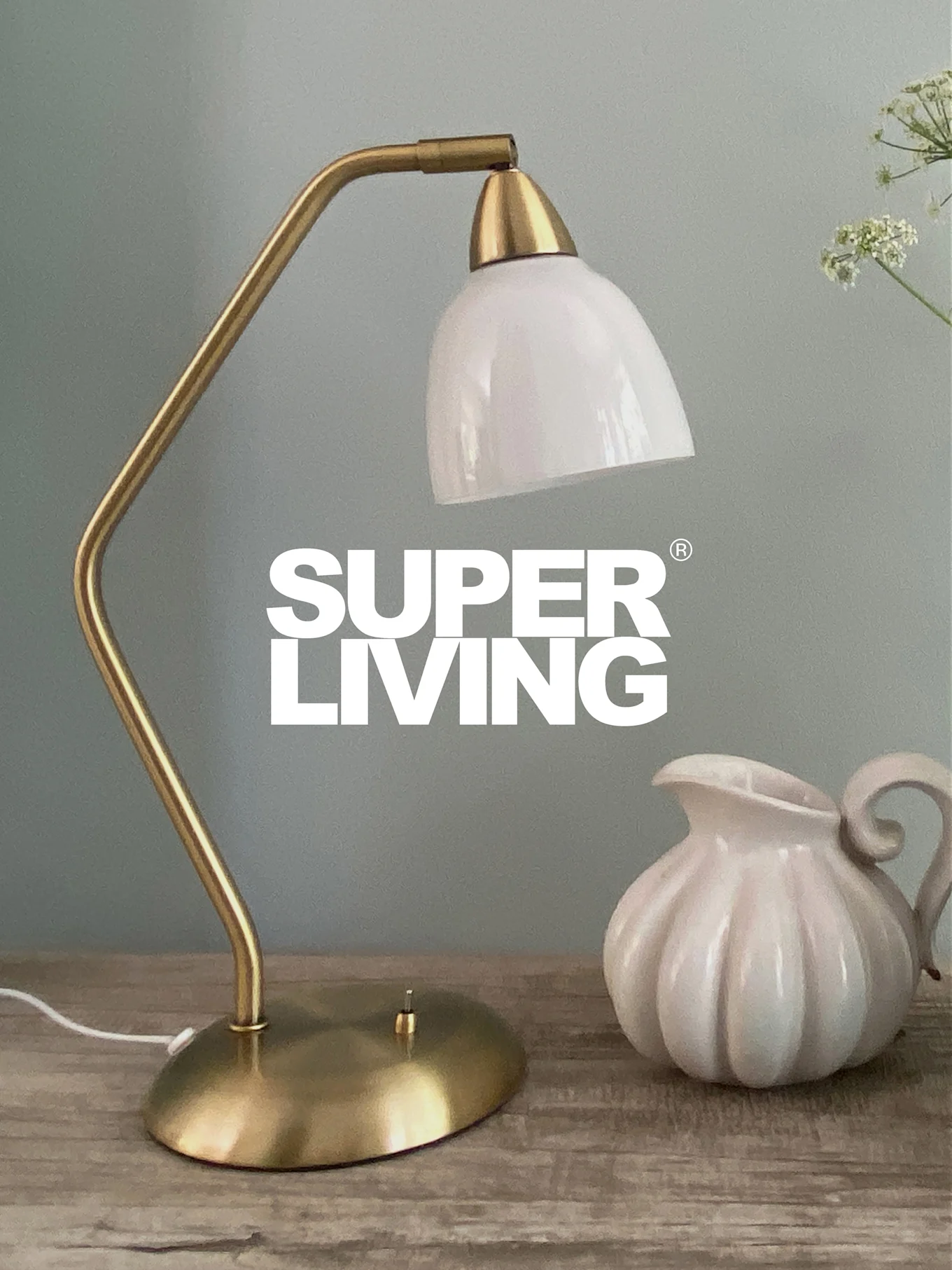 SUPER LIVING-branding over een messing bureaulamp met witte kap en een witte, geribbelde kan op een houten tafel.